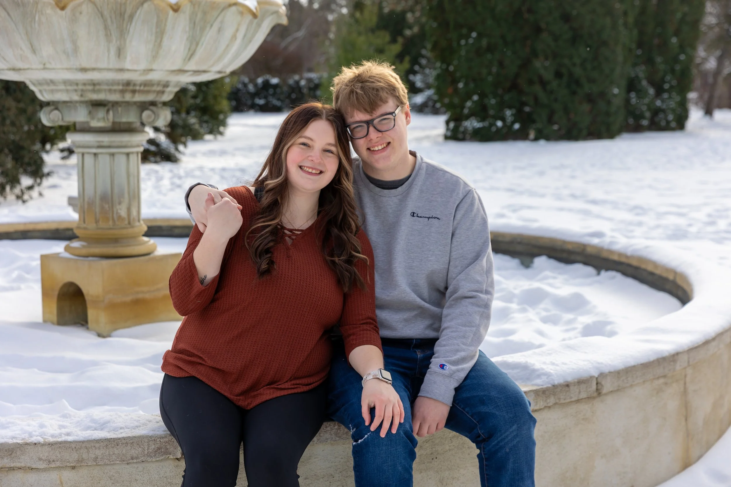 Minneapolis couples photography - A couple sit on the edge of a fountain next to each other with their arms around themselves in a winter scene at Arneson Acres Park in Edina, MN.