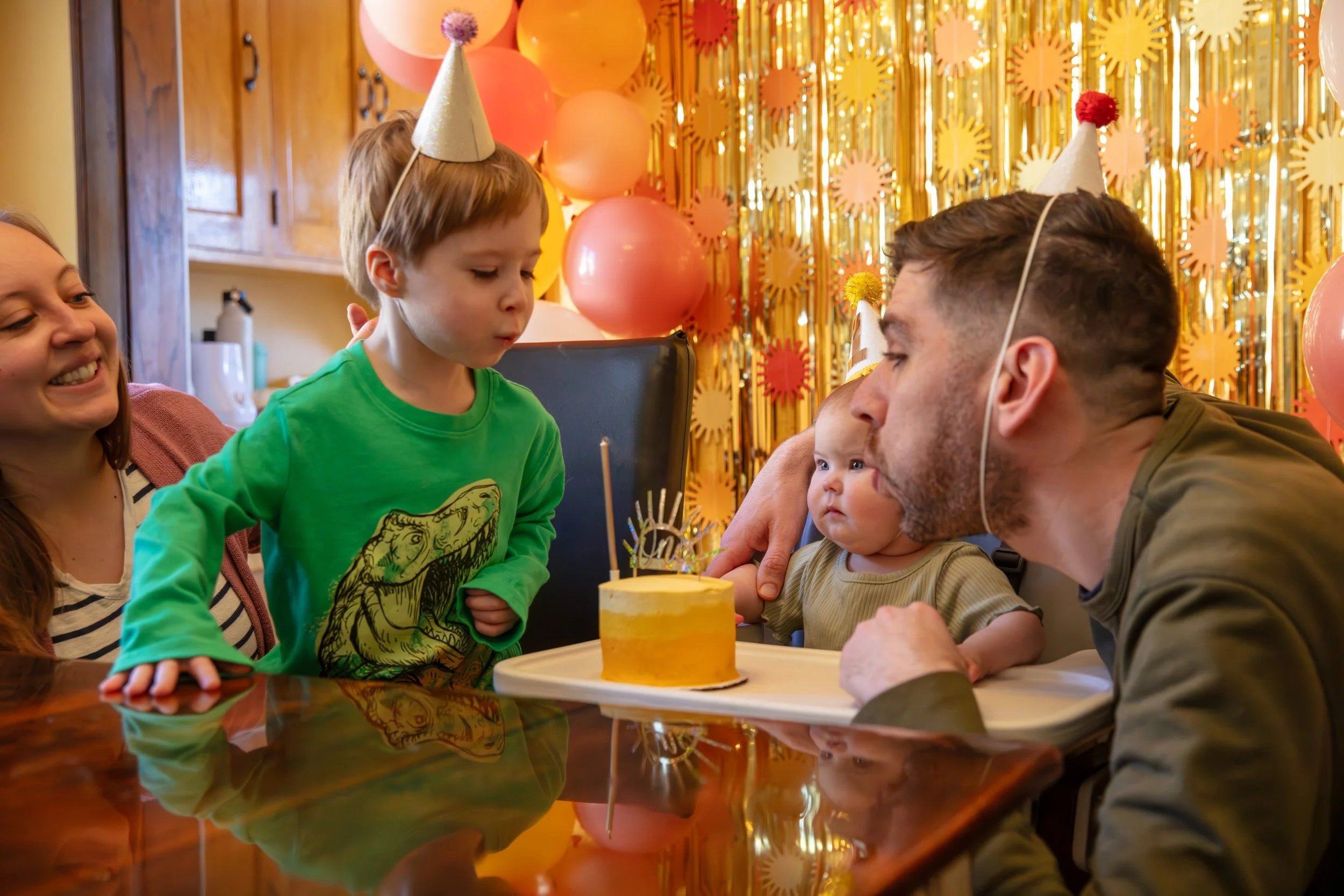 Minneapolis Cake Smash photographer - photo of a one year old baby in her highchair with a yellow frosted cake on the tray. She sits in front of birthday streamer and balloon decorations. Her father is on the right side of her, with her brother and m