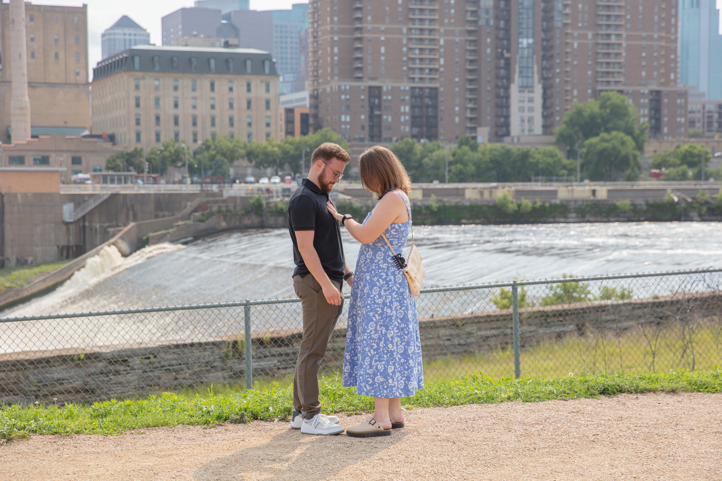 A couple, a man and woman, look at the woman's ring hand between them while on a park path right after he proposed to her in front of the falls and skyline at Water Power Park on St. Anthony Main.