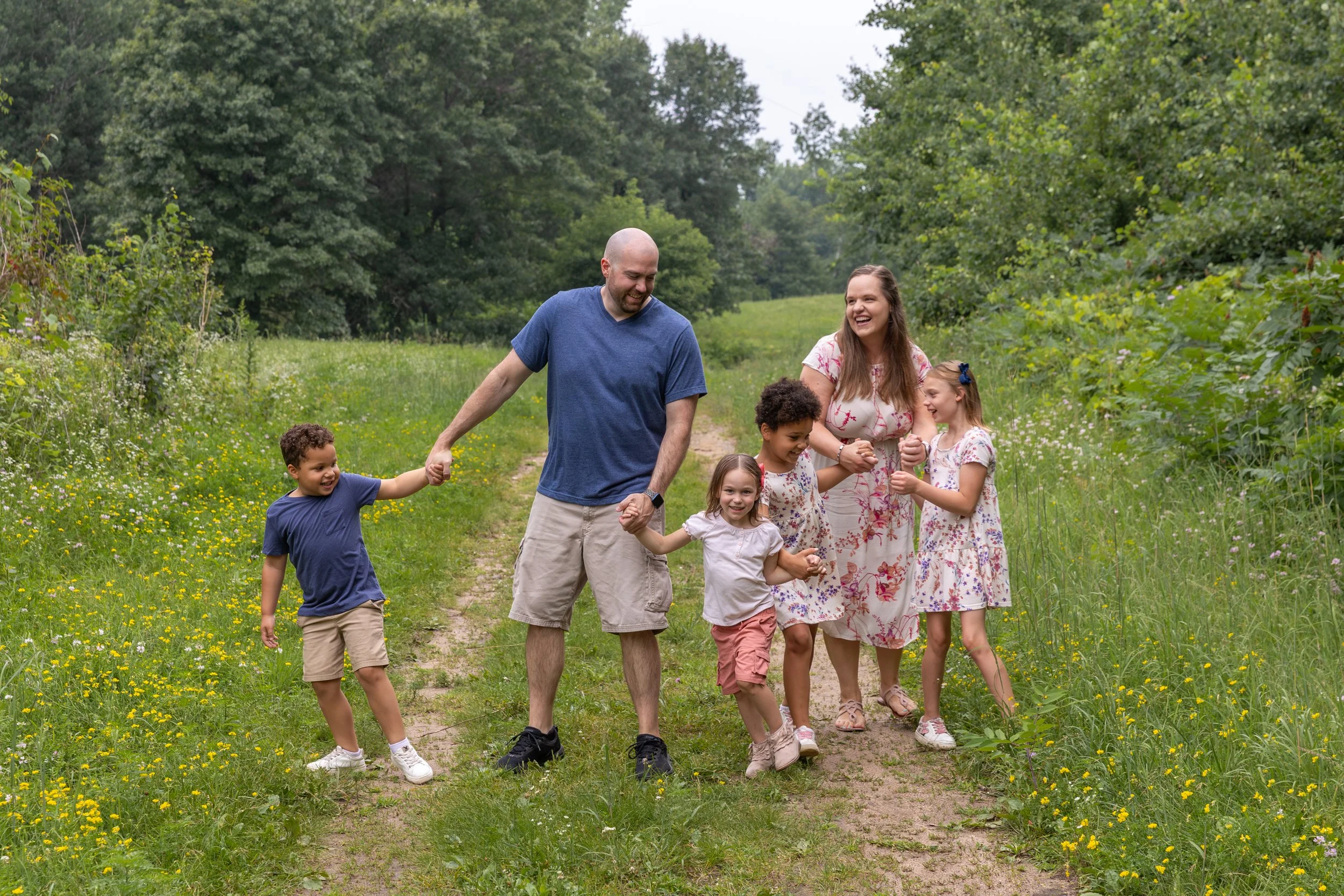 A blended family of a dad with his two daughters and a mother with her daughter and son all walk hand in hand together bumping into each other laughing as they walk along a trail in Eagan, MN.