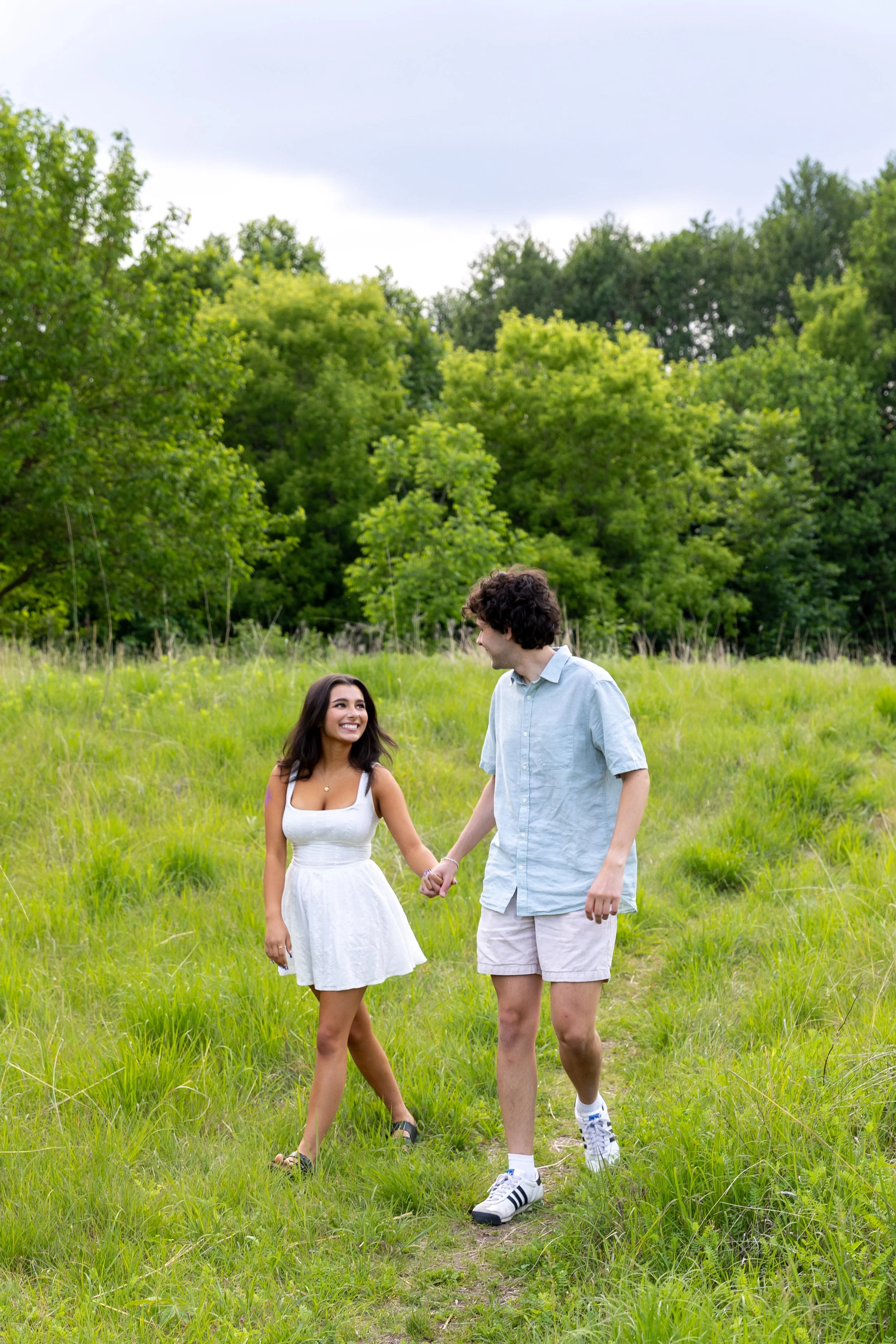 Minneapolis couples photography - A couple holds hands while walking towards the camera in a grassy field looking at each other and smiling while at Minnehaha Falls Park in Minneapolis, MN. 