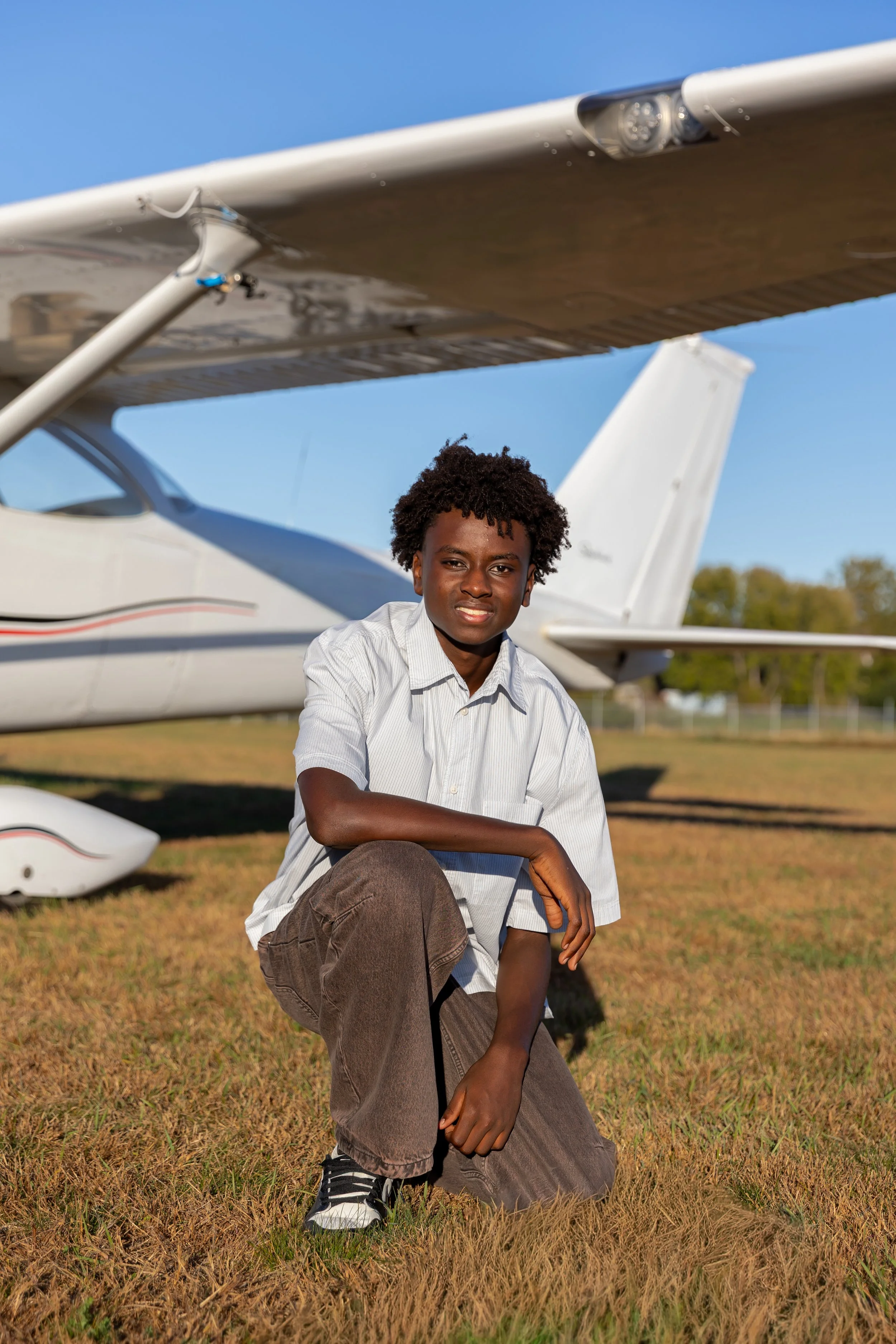 A Maple Grove high school senior squats in front of a Cessna airplane in the Crystal airport smiling at the camera for their senior photos.