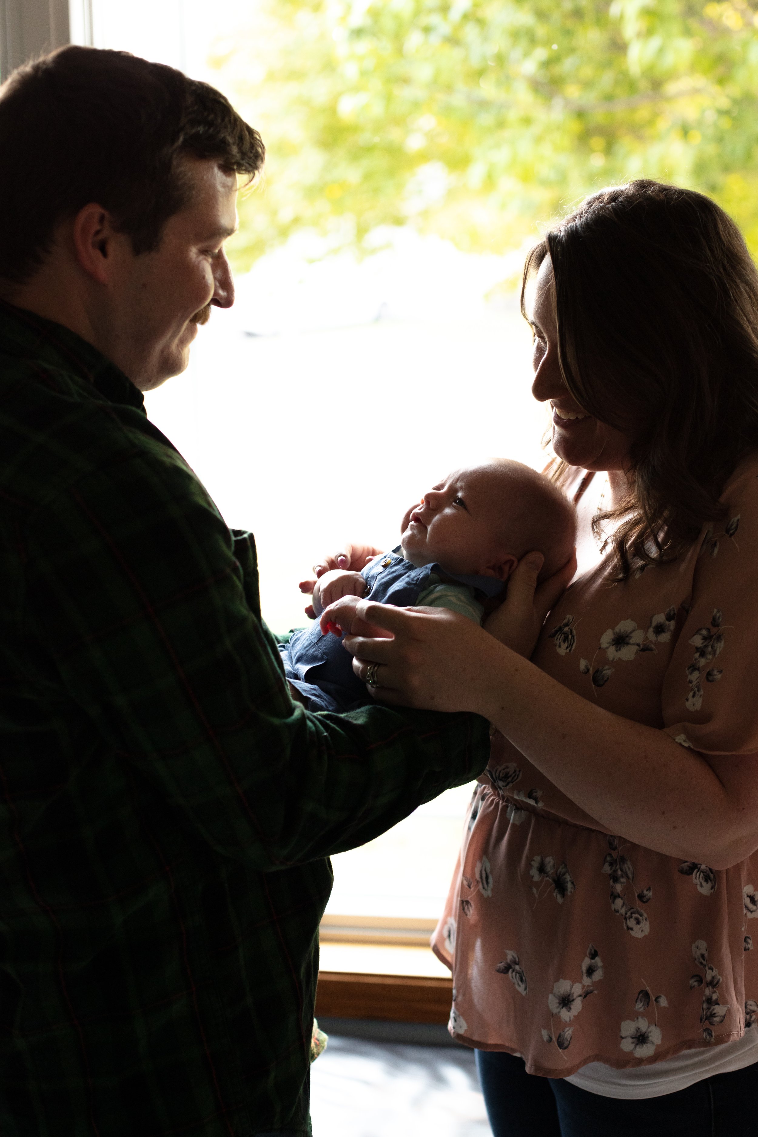 Silhouette of dad holding their baby cradling it's head in his hands while mom stands on the other side with her hands wrapped around the baby as well. Both mom and dad gaze down at their baby.