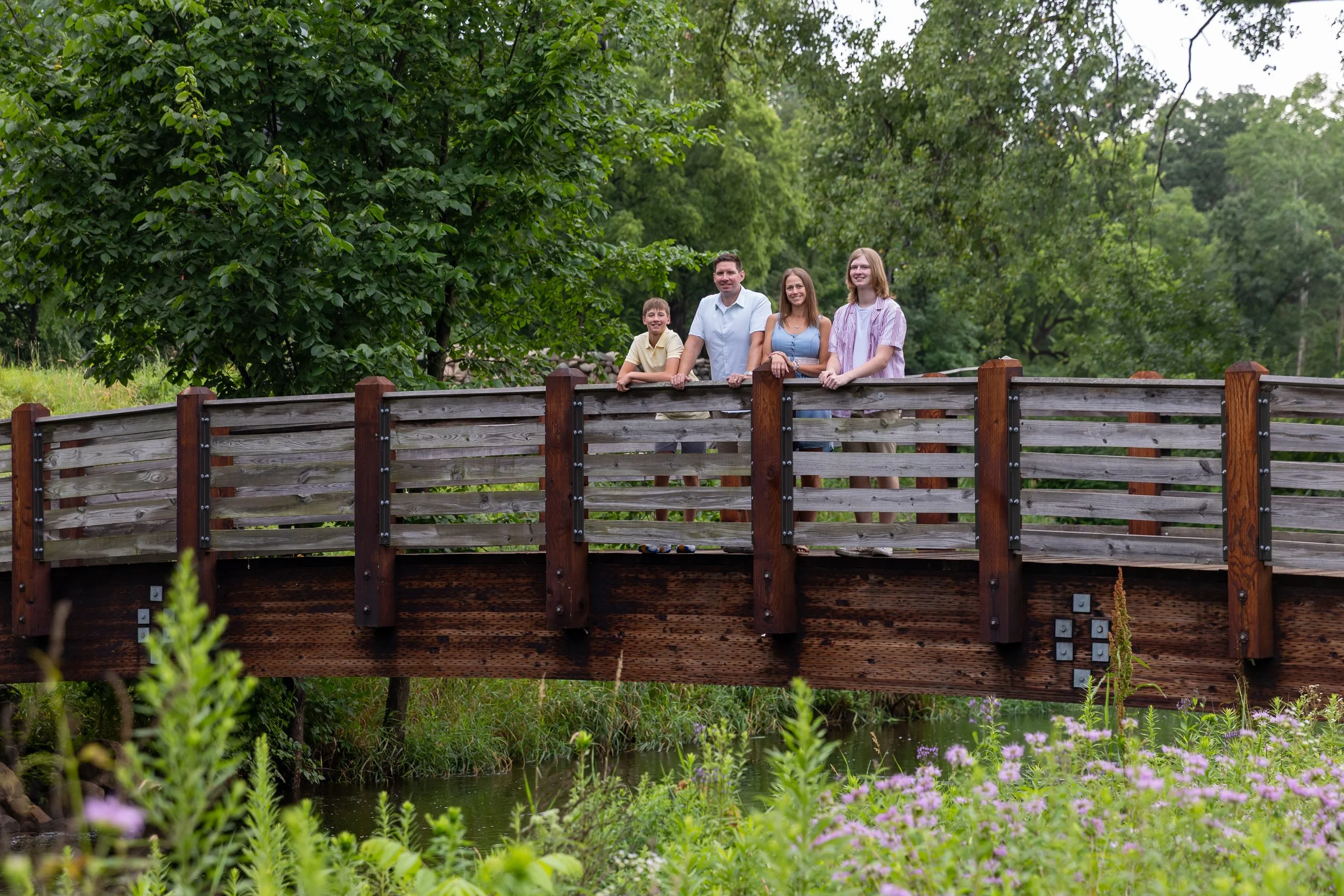 A family of four - a father and mother stand in the middle of their two teenage sons - on a bridge above a creek at Minnetonka Mills Park in Hopkins, MN smiling at the camera. 