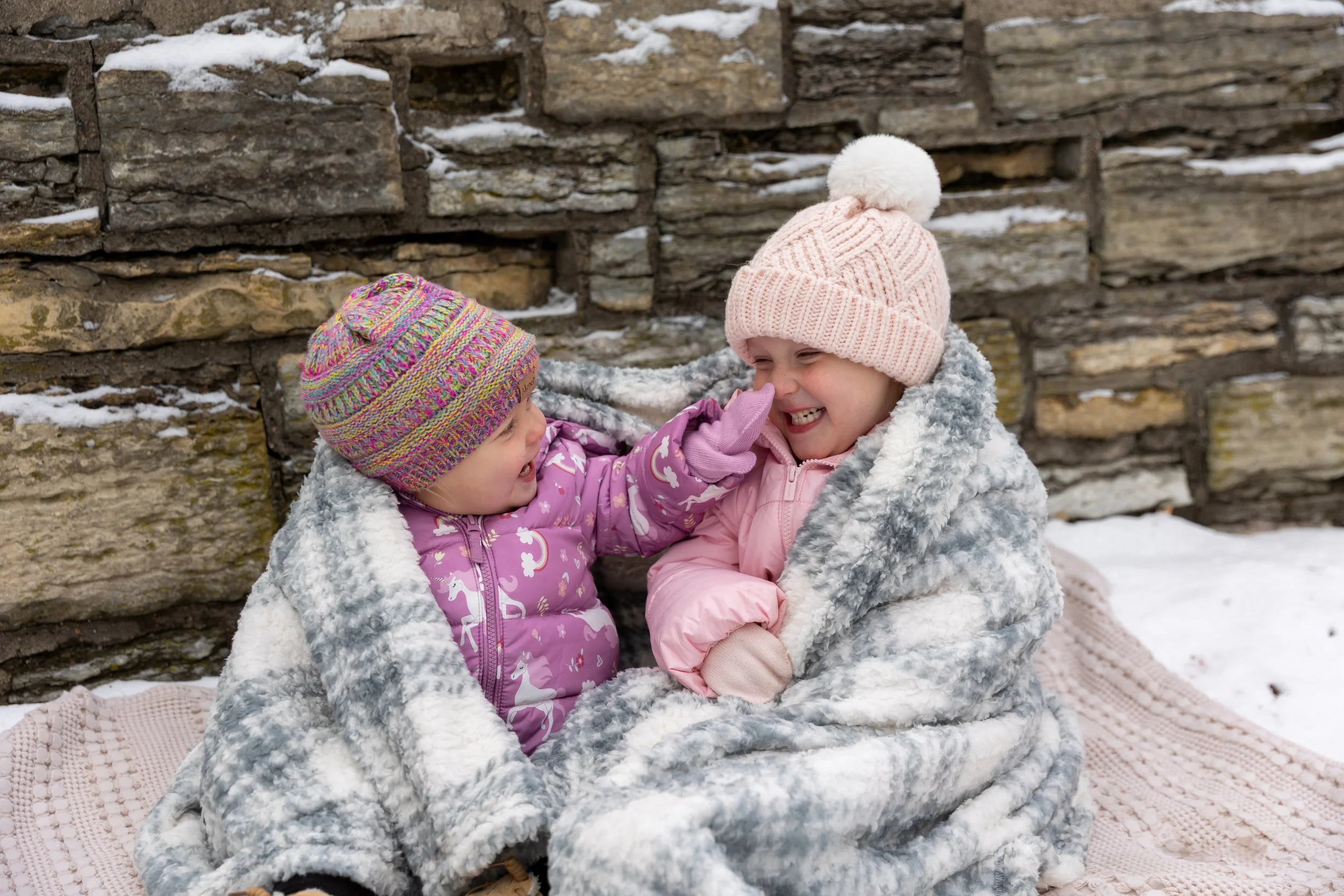Minneapolis family photography - Two young toddler girls are sitting all bundled up in winter jackets and a blanket while smiling at each other booping each other on the nose laughing. It is a snowy winter scene at Minnehaha Falls in Minneapolis, MN.