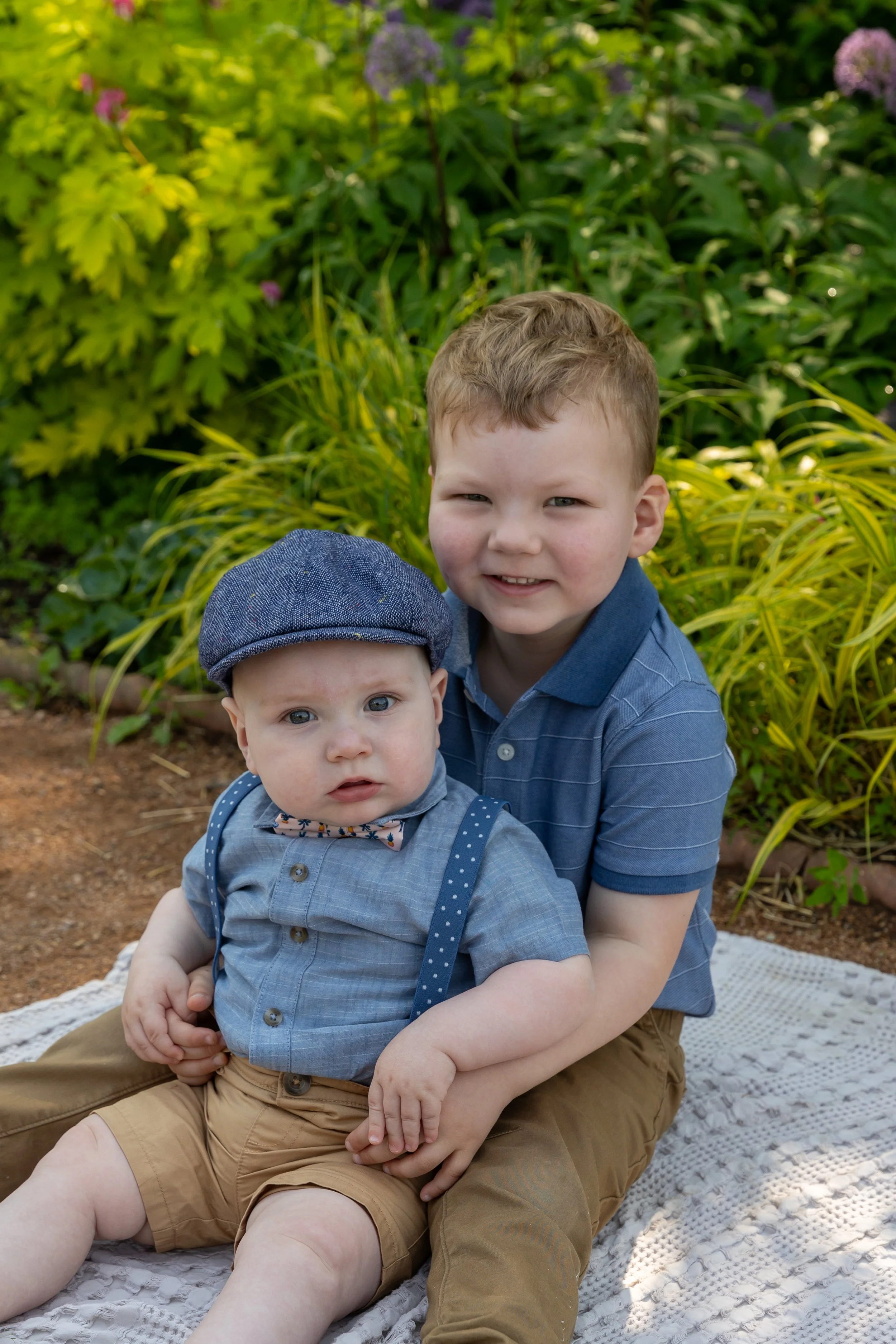 Minneapolis family photography May Mini Session- Two brothers, a toddler and baby, sit on a blanket together smiling at the camera. They are at Lyndale Rose Garden in Minneapolis, MN.
