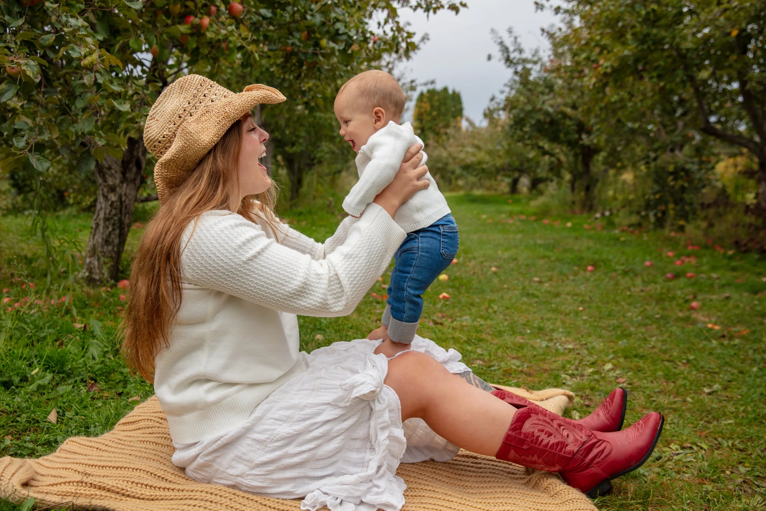 A mother sits on a blanket in the middle of Aamodt's Apple Orchard while holding her baby facing her as they smile at each other.