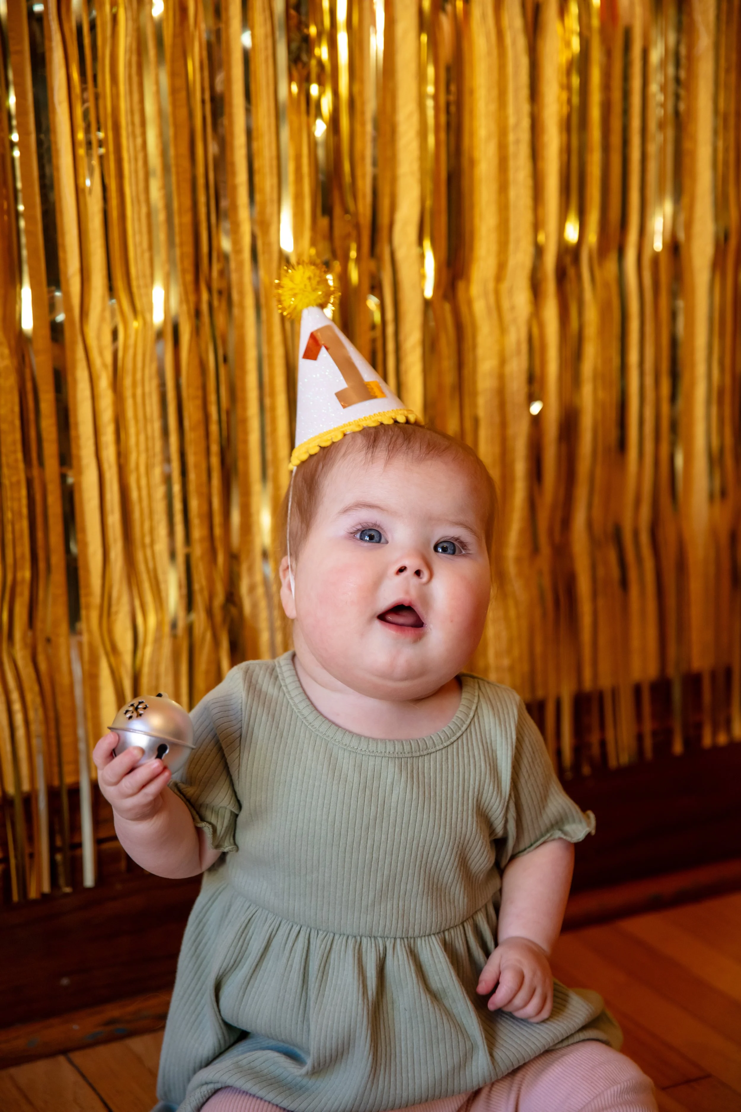 Minneapolis Cake Smash photographer - photo of a one year old baby is sitting on the floor looking up at the camera and is sitting in front of birthday streamers.  She is wearing a green dress and a birthday hat with the number 1 on it.