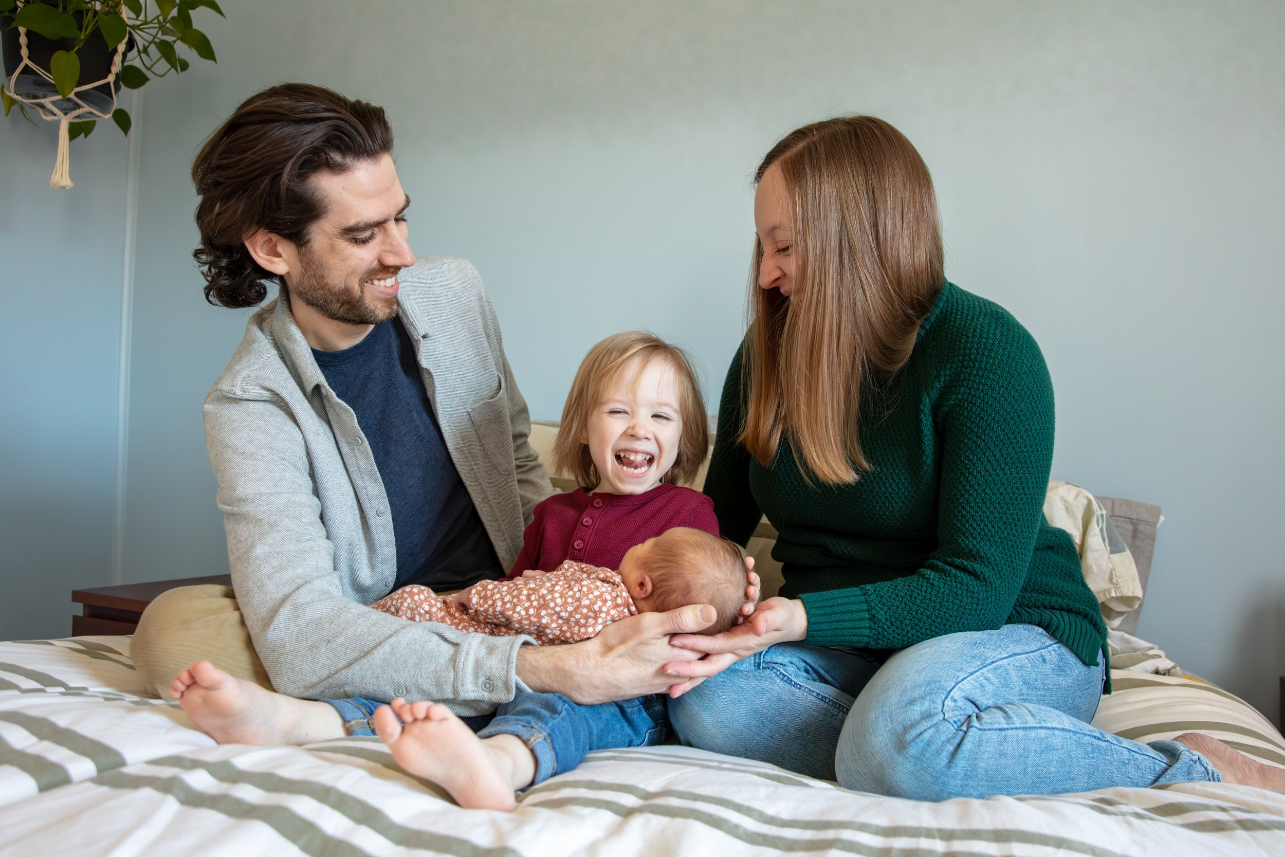 A father and mother sit on their bed with their toddler between them laughing as he holds his newborn sister.