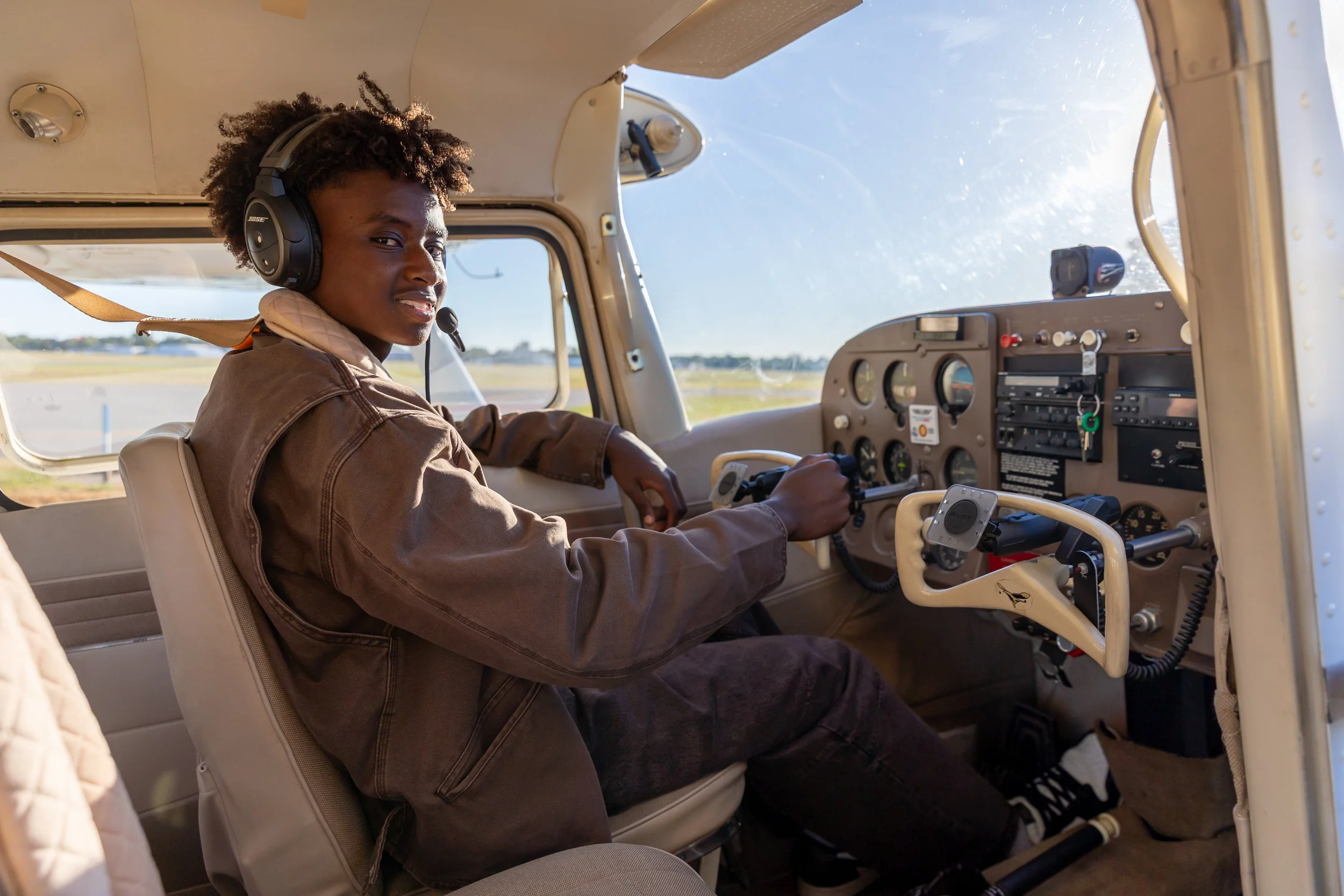 A Maple Grove high school sits in the cockpit at the controls of a Cessna airplane in the Crystal airport smiling at the camera for their senior photos while wearing a headset.
