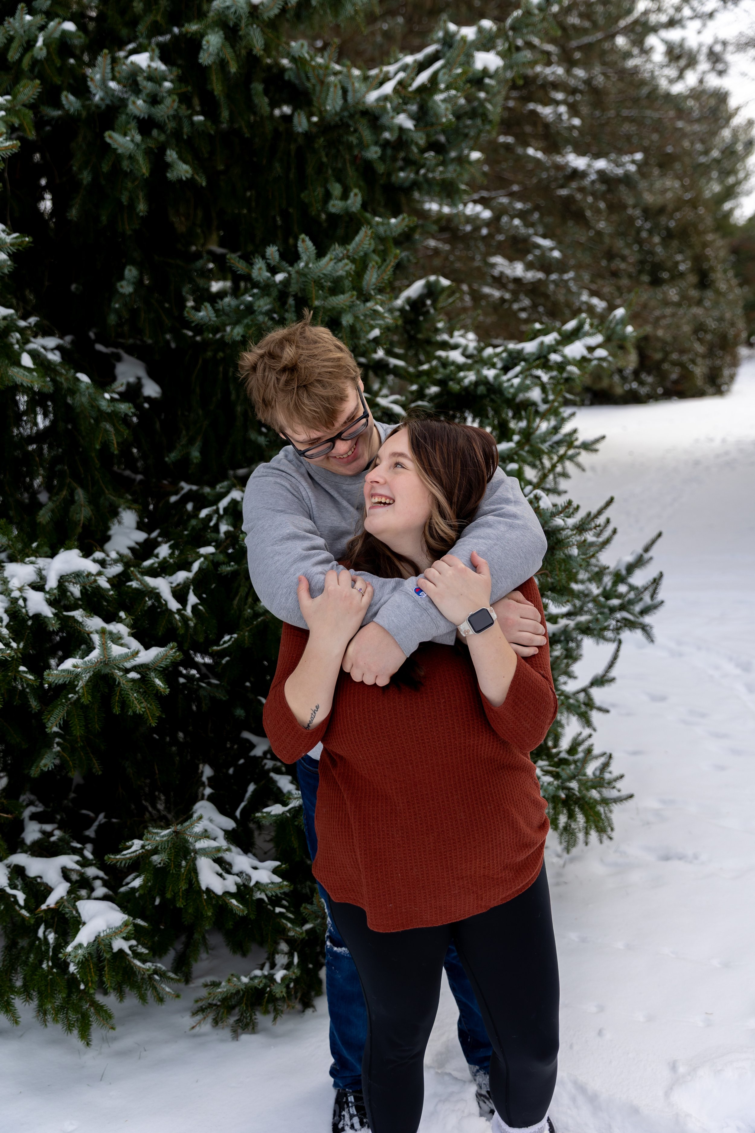 Minneapolis couples photography - A couple embrace and smile at each other with the man looping his arms around her from behind in a winter scene at Arneson Acres Park in Edina, MN.