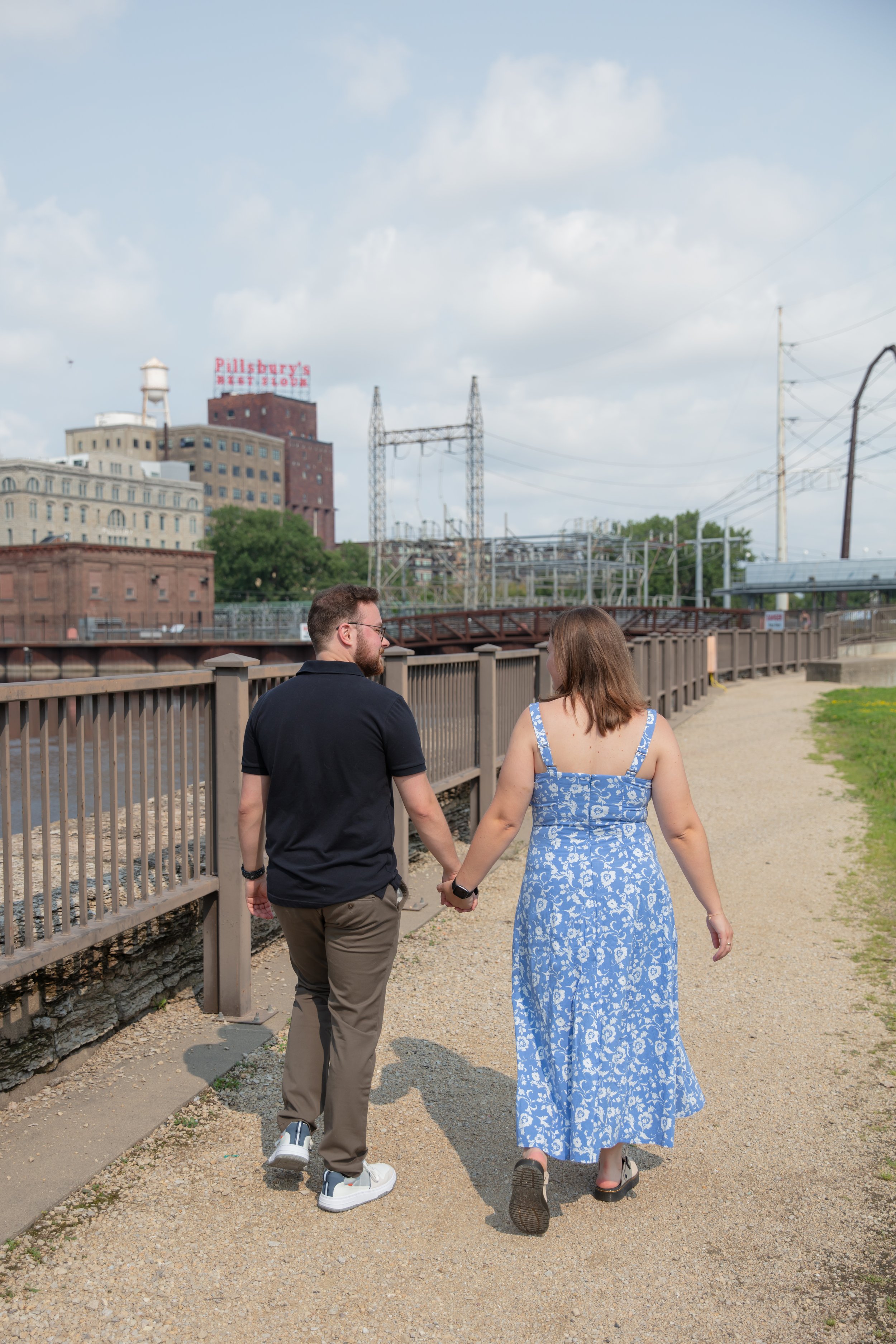 A couple, a man and woman, walk away from the photographer holding hands smiling at each other after he proposed to her in front of the falls and skyline at Water Power Park on St. Anthony Main.