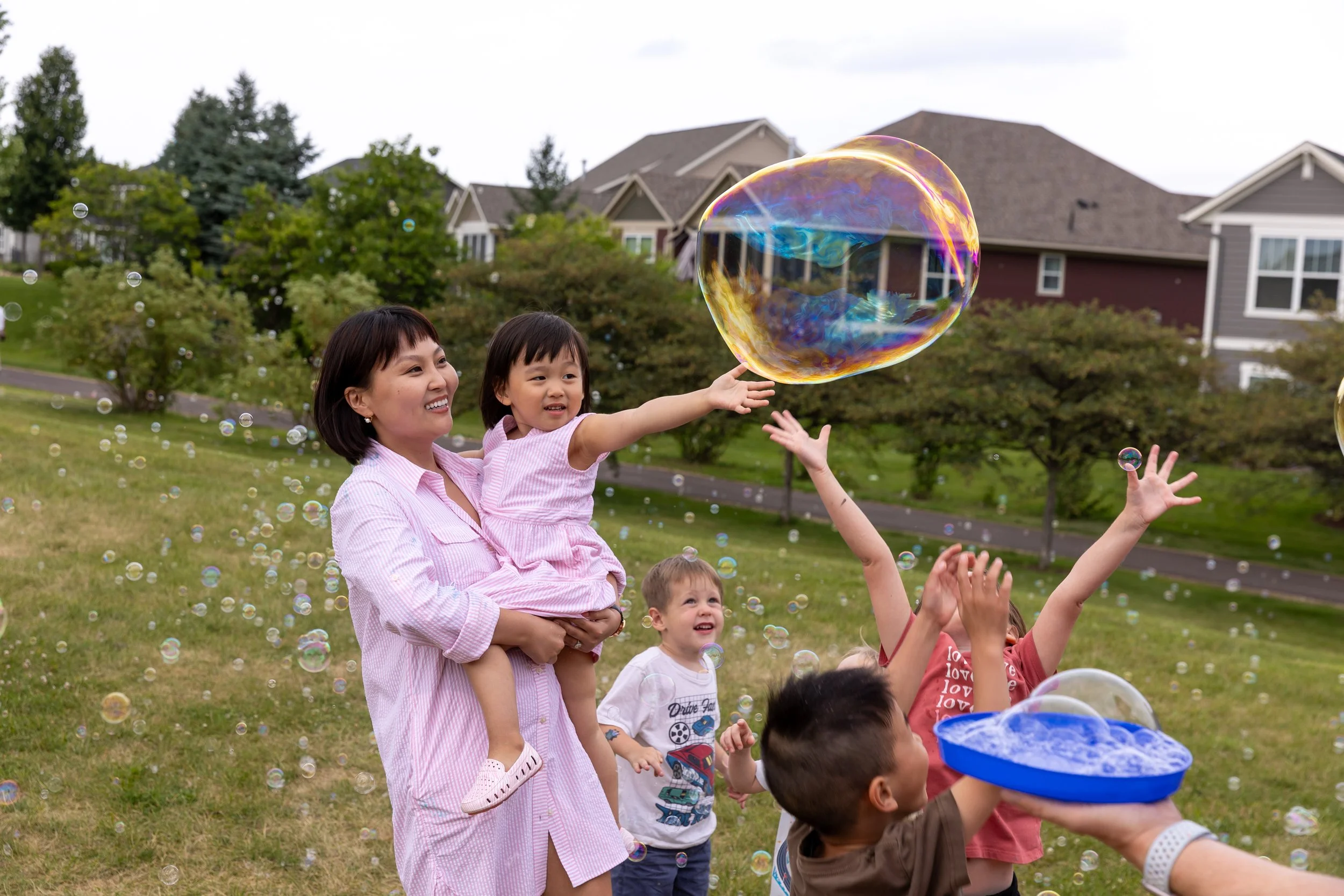 A mom in a pink dress smiles while holding her daughter up, also wearing a pink dress, in a St. Paul Park with bubbles blowing around them from a bubble machine. Other children are running and playing around them in the bubbles.