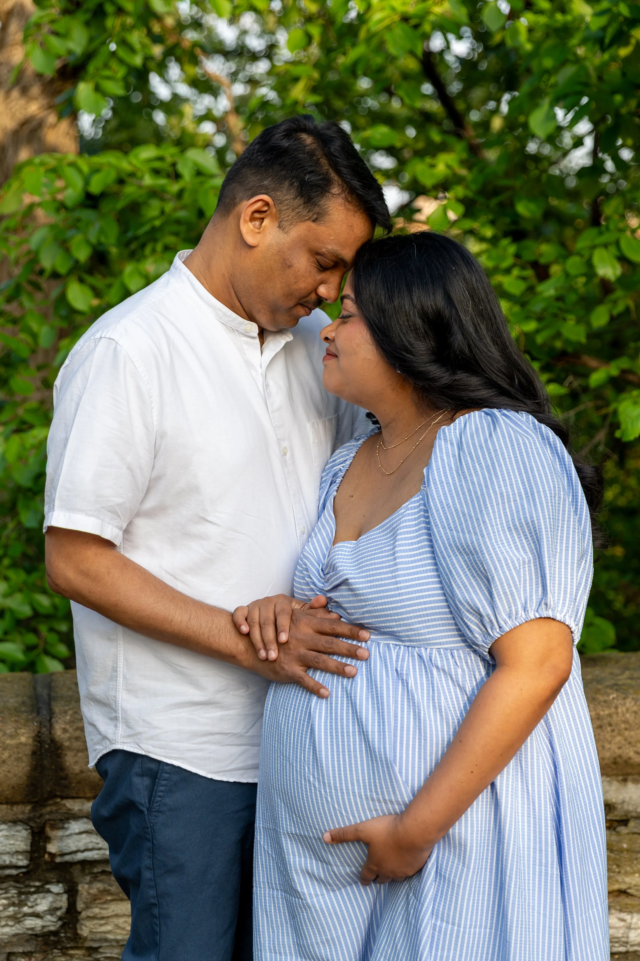 Maternity session - father and mother stand together while gazing lovingly at each other at Minnehaha Falls Park while holding the baby bump