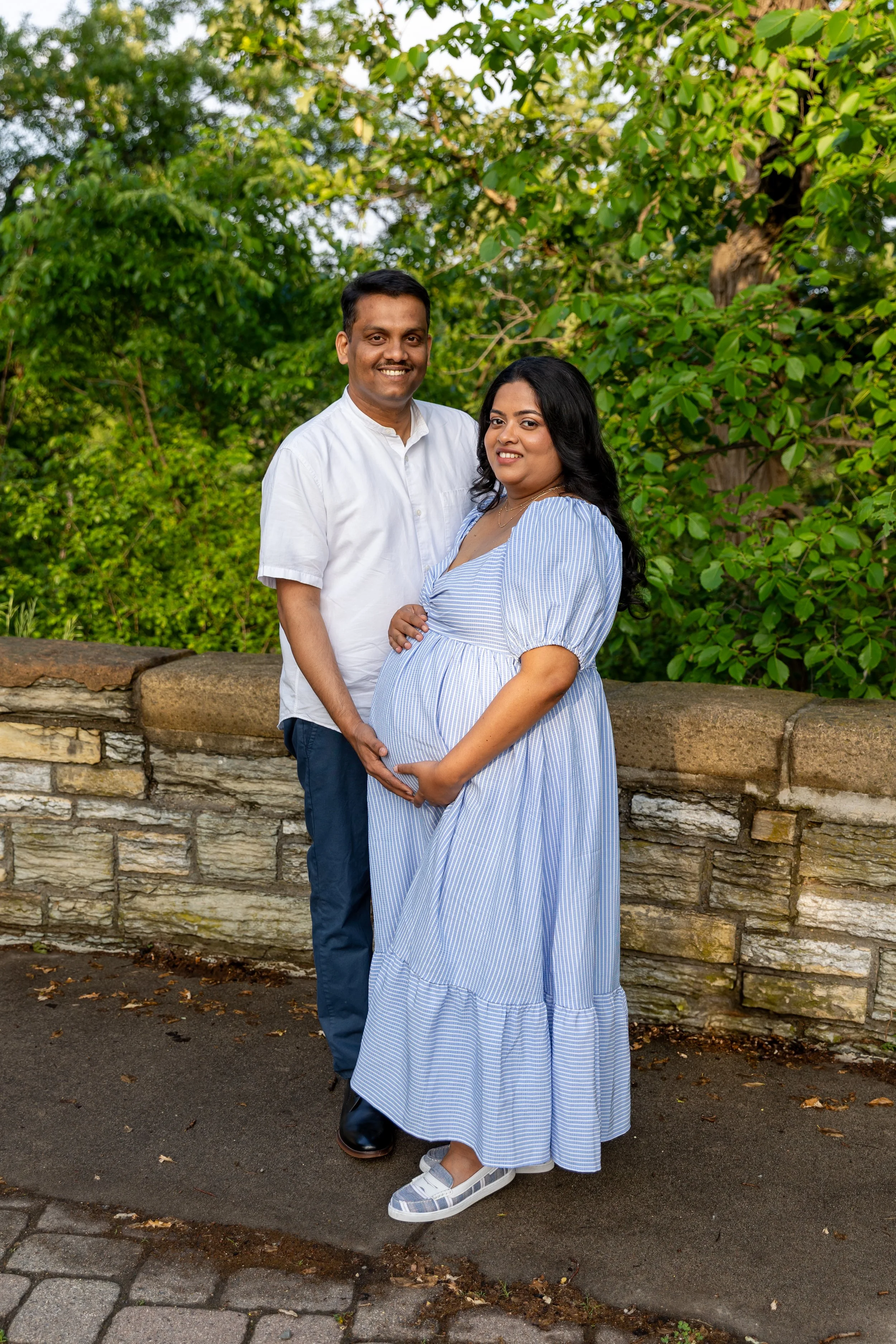 Maternity session - father and mother stand together smiling at the camera while cradling each other and the baby bump while at Minnehaha Falls Park 