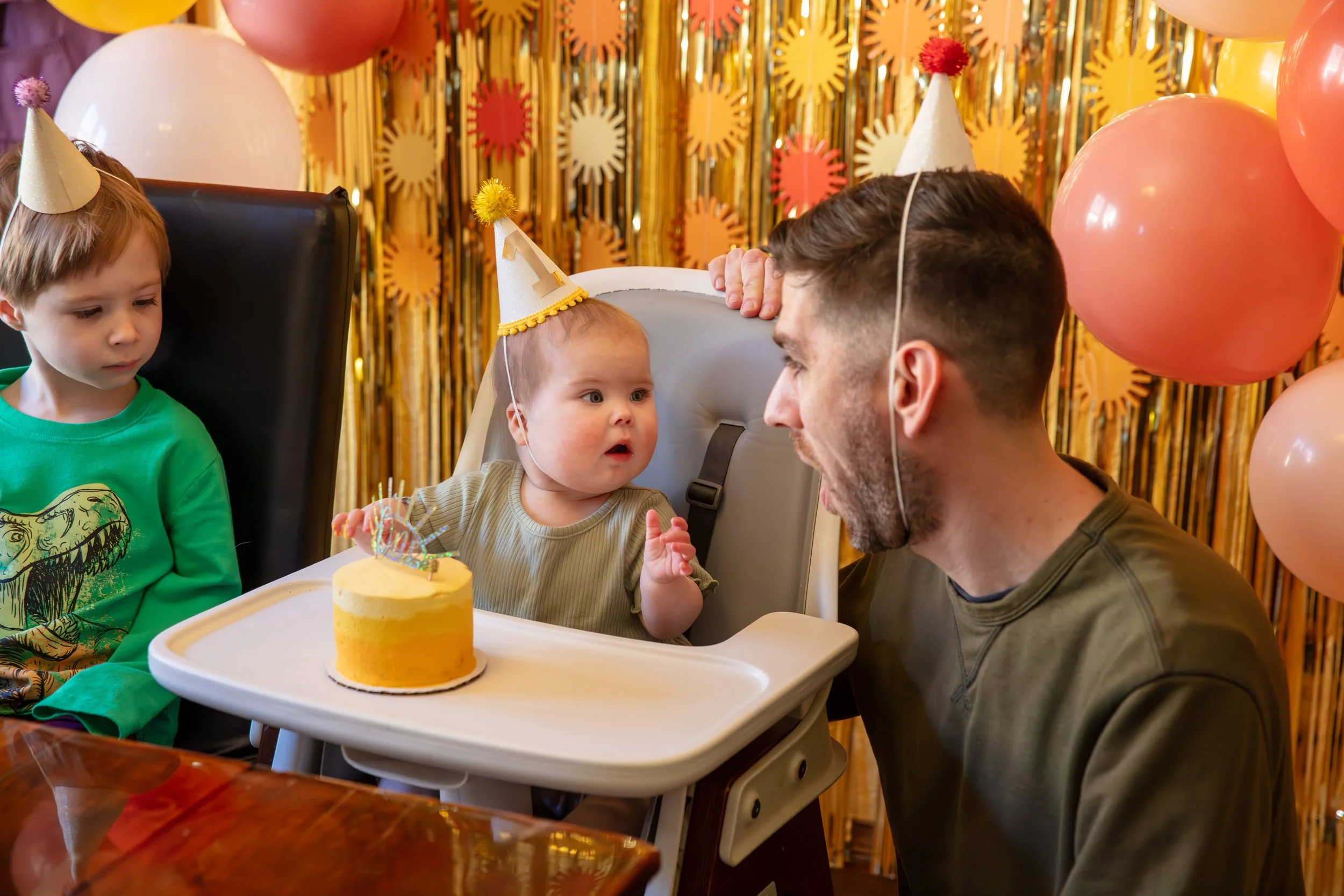 A one year old sits in her highchair with a birthday hat on while her father looks at her with a happy shocked face. Her yellow smash cake is sitting in front of her with her older brother looking at it longingly.