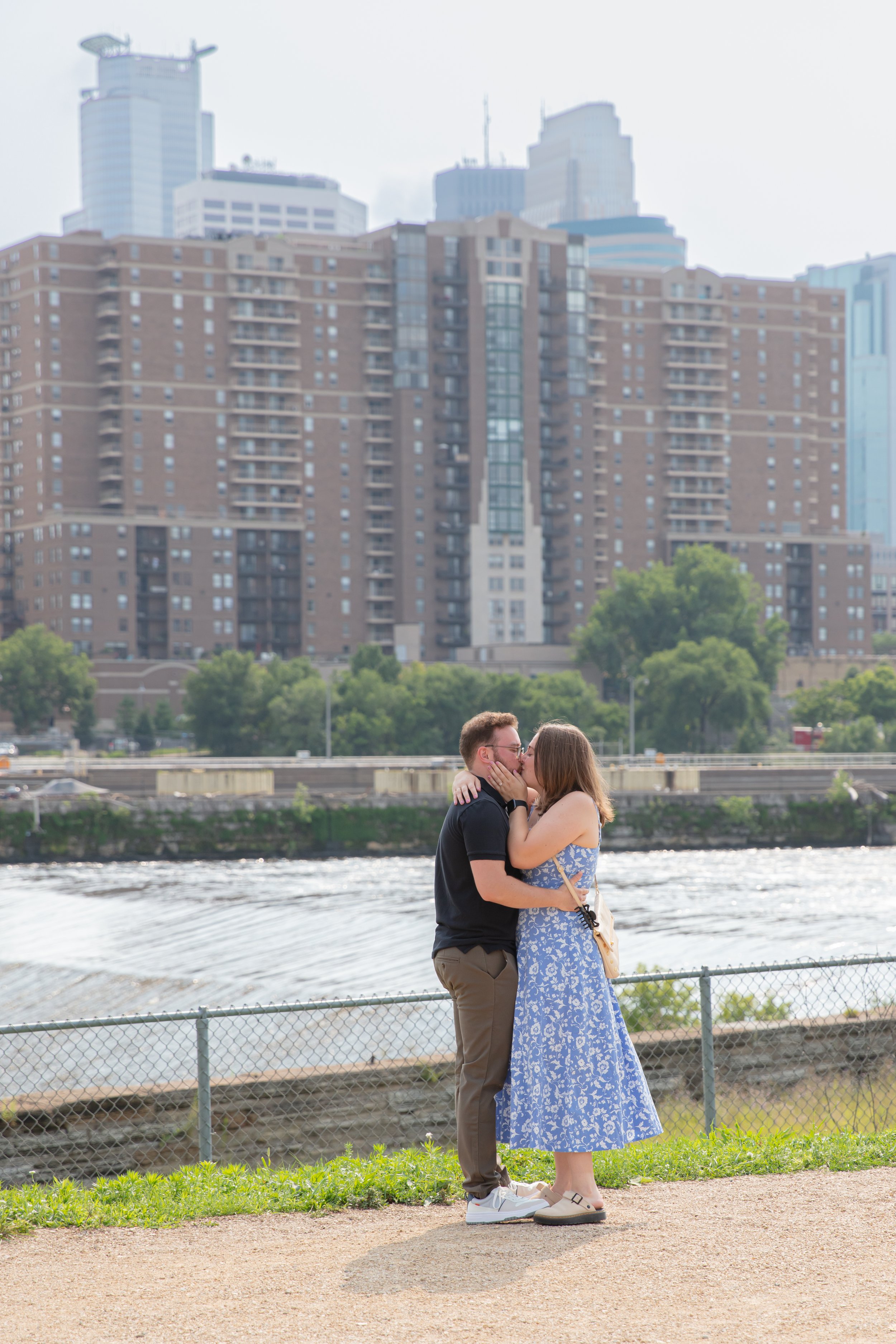 A couple, a man and woman, embraces each other kissing on a park path right after he proposed to her in front of the falls and skyline at Water Power Park on St. Anthony Main.