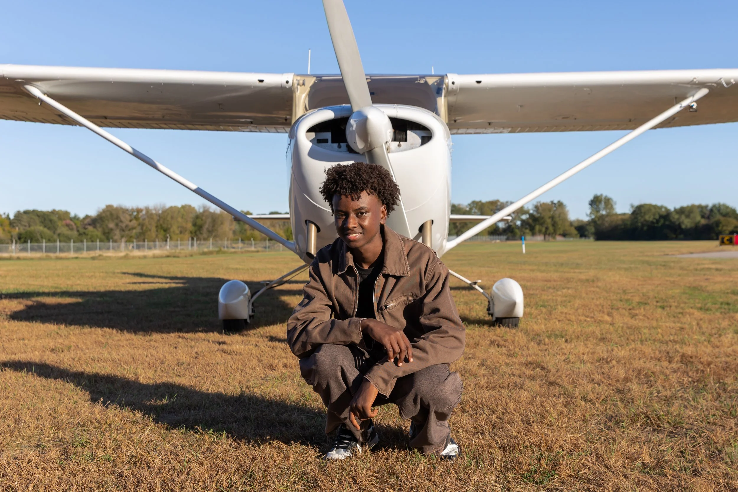 A Maple Grove high school senior squats in front of a Cessna airplane in the Crystal airport smiling at the camera for their senior photos.