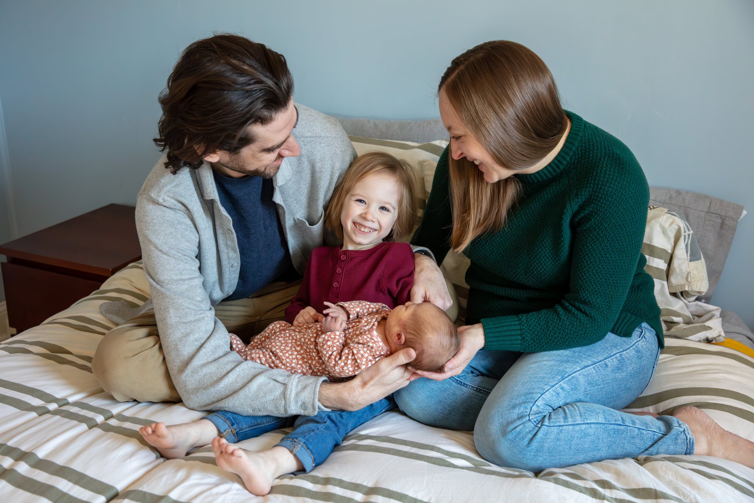 A father and mother sit on their bed tickling their toddler between them as he holds his newborn sister.