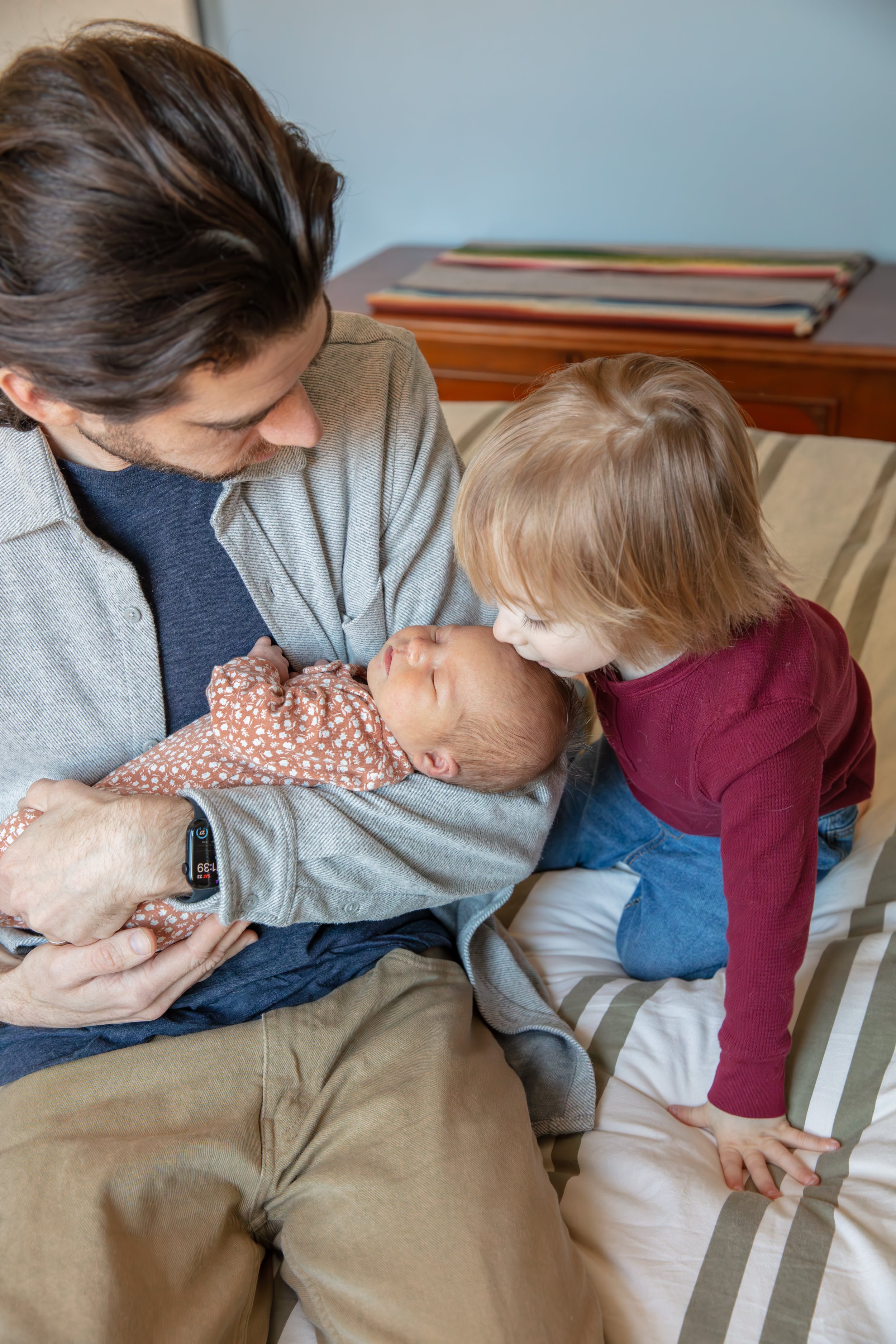 Father holds the newborn in his arms while his toddler sits next to them and kisses the baby on the forehead.