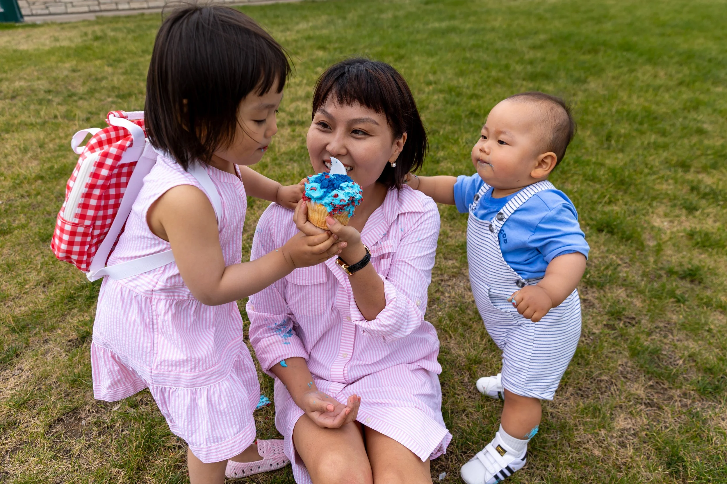 A mom in a pink dress sits in the grass in a St. Paul park between her daughter and son, smiling at her kids while holding a cupcake up. Her daughter is standing next to her in a pink dress with a red backpack on reaching for cupcake. Her son is hold