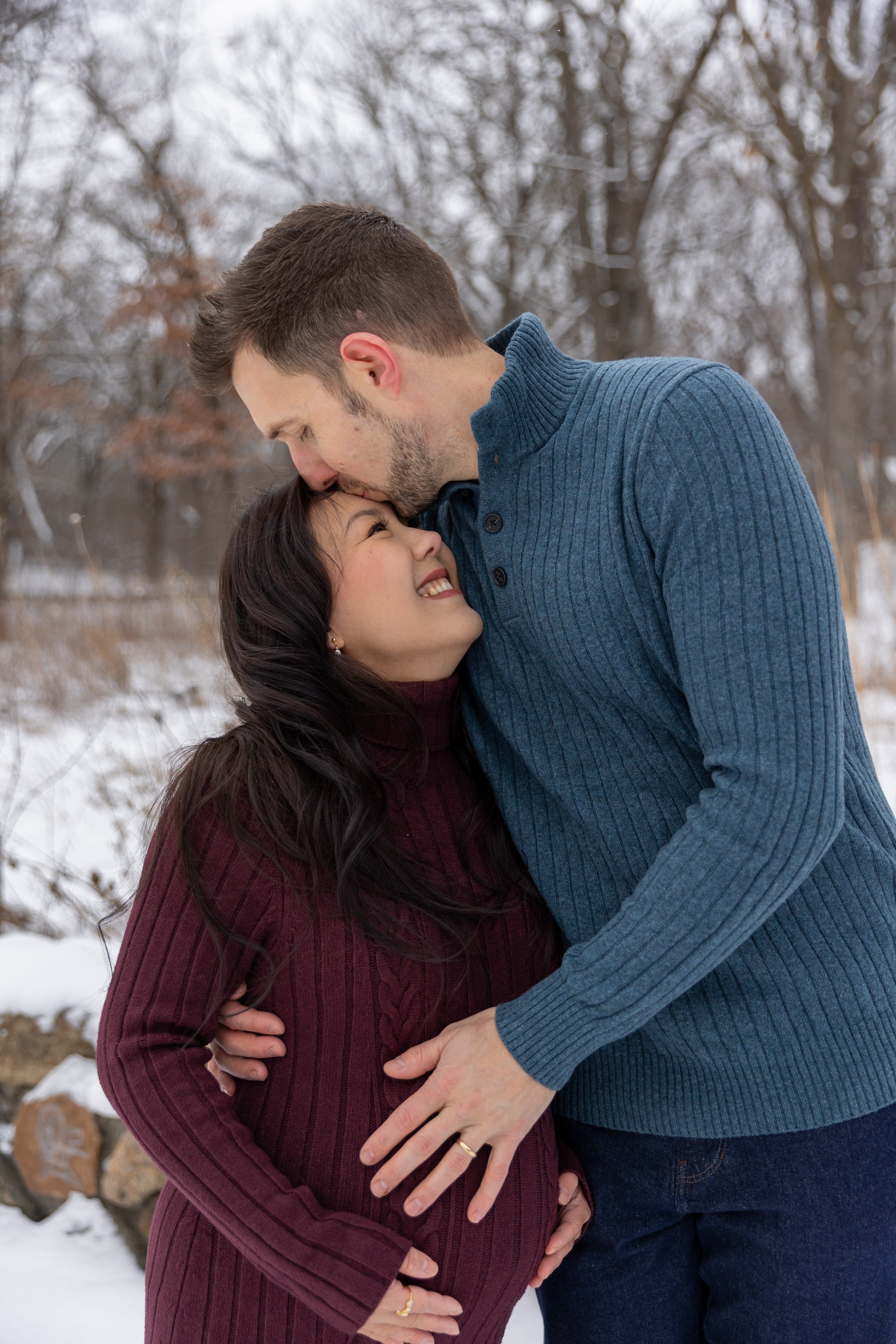 Minneapolis Maternity photography session - A soon to be mother and father stand embracing each other side by side while the father kisses the forehead of the mother at Minnetonka Mills Park in Hopkins, MN in the winter.