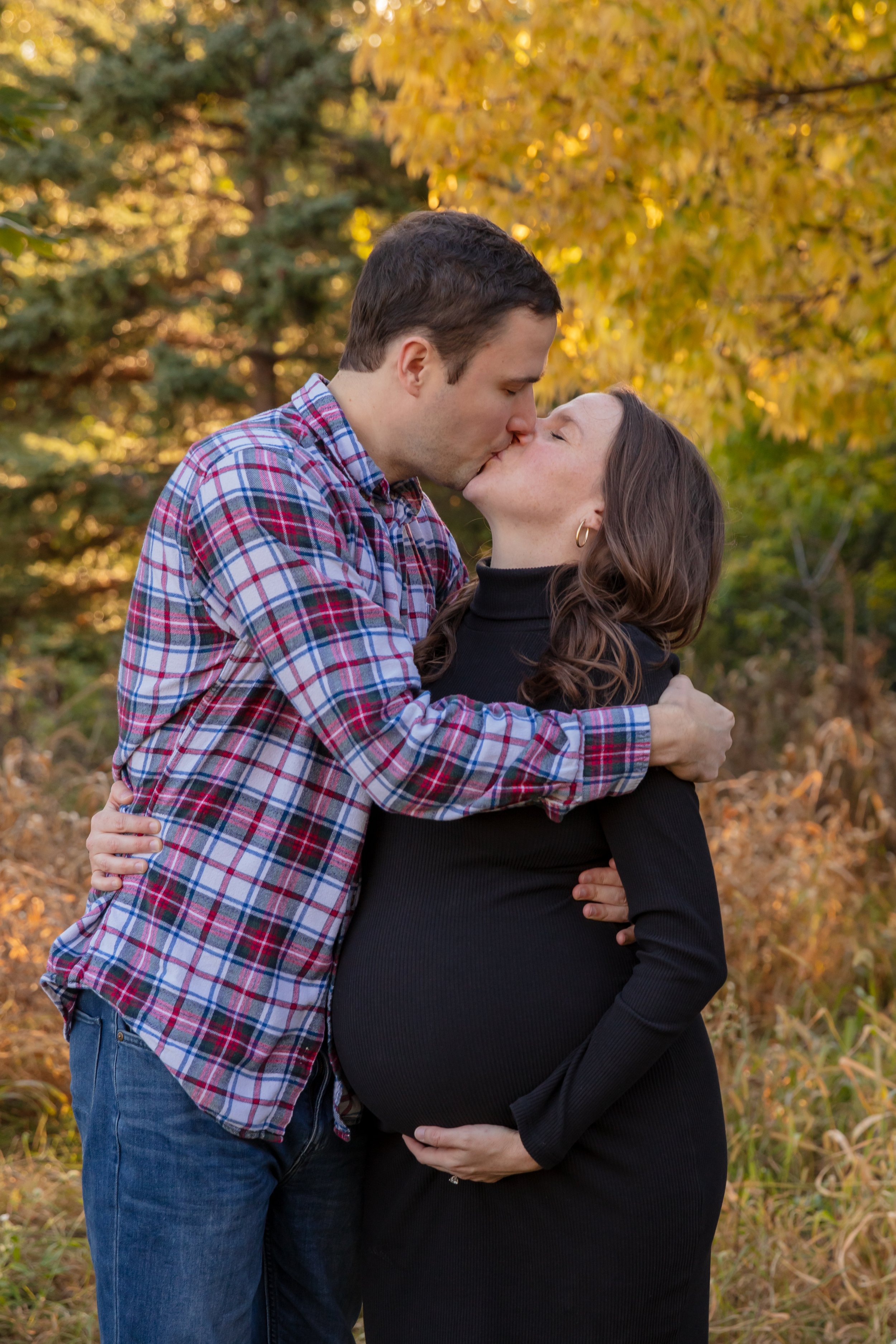 Maternity session, expecting mother and father-to-be kiss each other with their arms around each other in a Minneapolis park with fall foliage behind them.