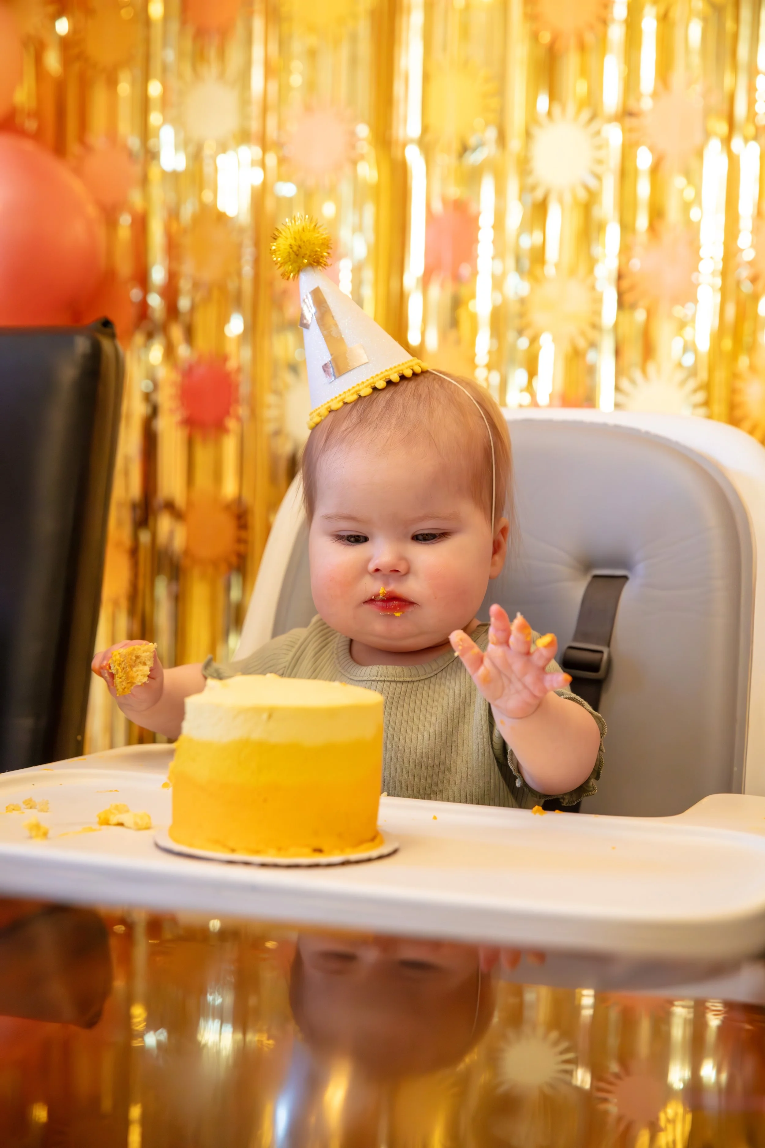 Minneapolis Cake Smash photographer - photo of a one year old baby in her highchair with a yellow frosted cake on the tray. She sits in front of birthday streamer and balloon decorations.  She is about to stick her hands in the frosting of the cake.