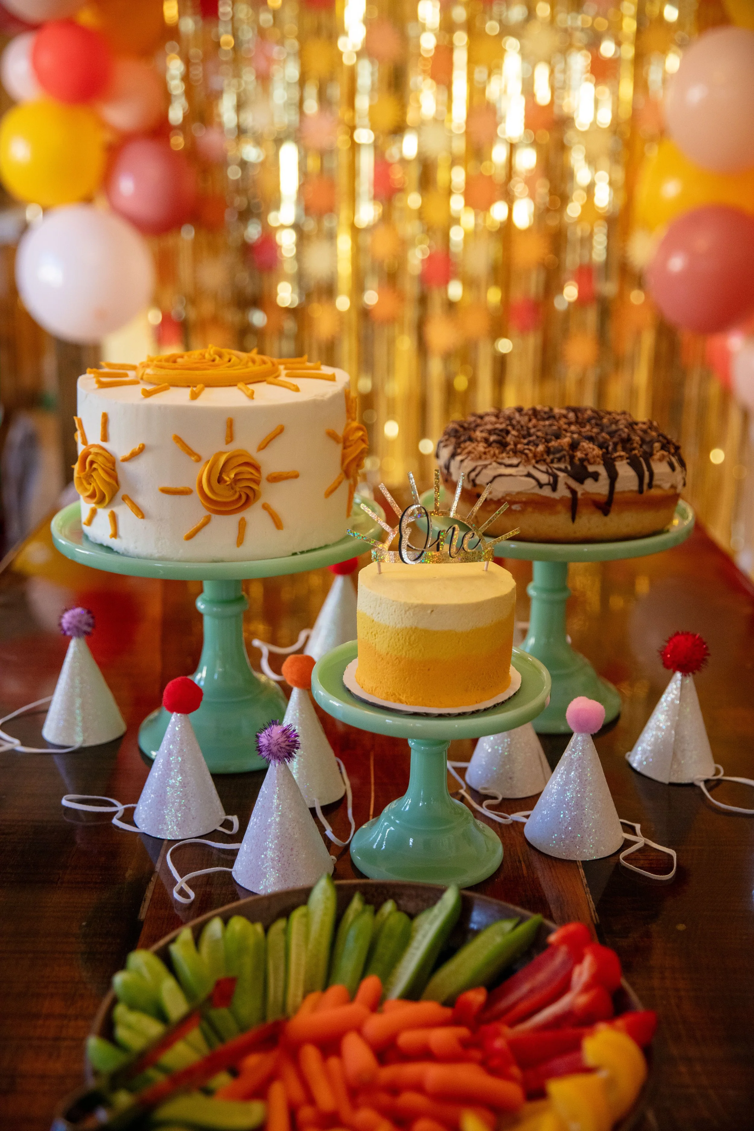 Minneapolis cake smash photographer - photo of three birthday cakes in front of birthday streamers, birthday hats and decorations. The smallest cake is decorated in yellow frosting with a cursive "One" cake topper on top.