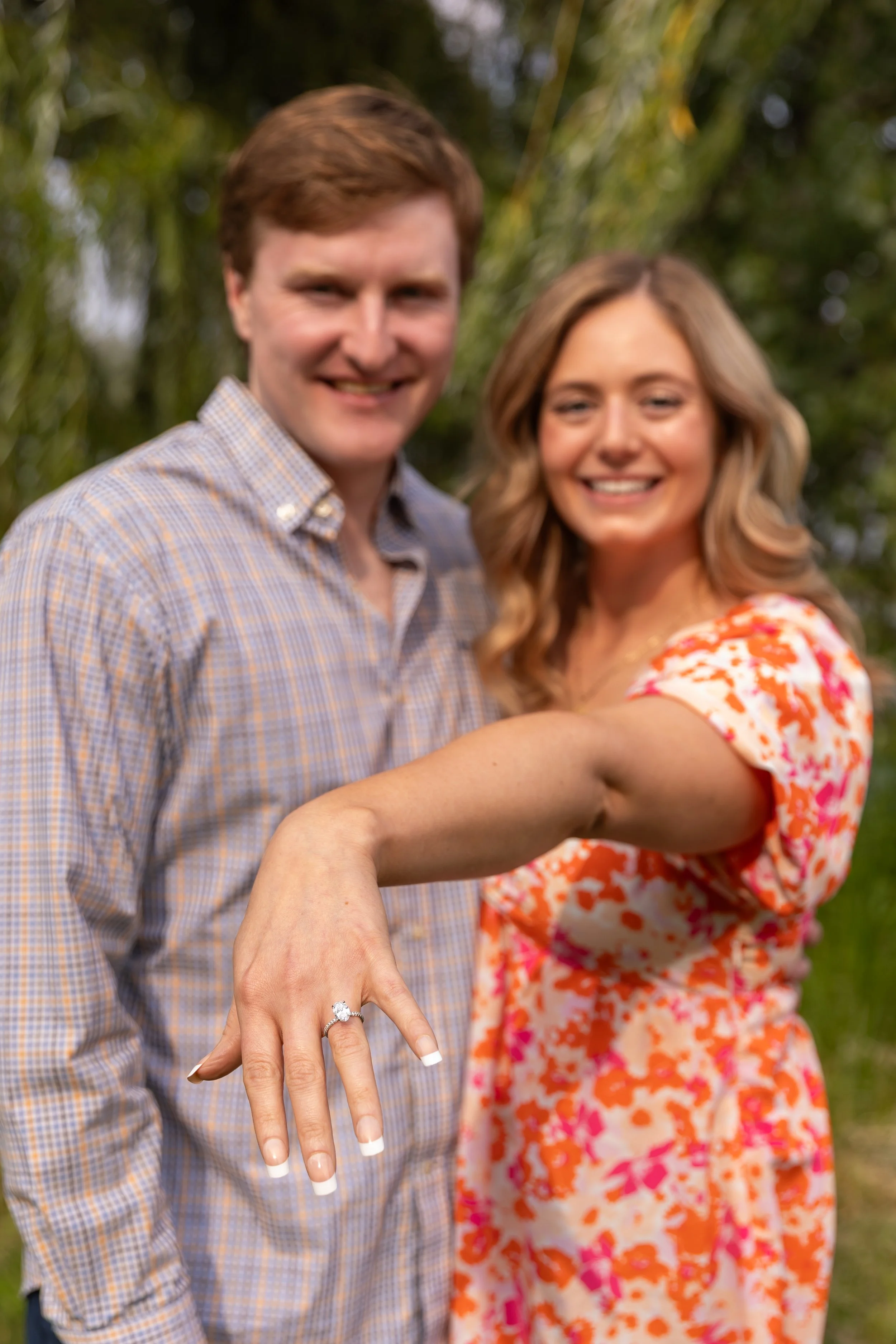 Minneapolis surprise proposal photography - A man and woman stand cuddled together smiling at the camera while she shows off her new engagement ring at Lake Harriet.