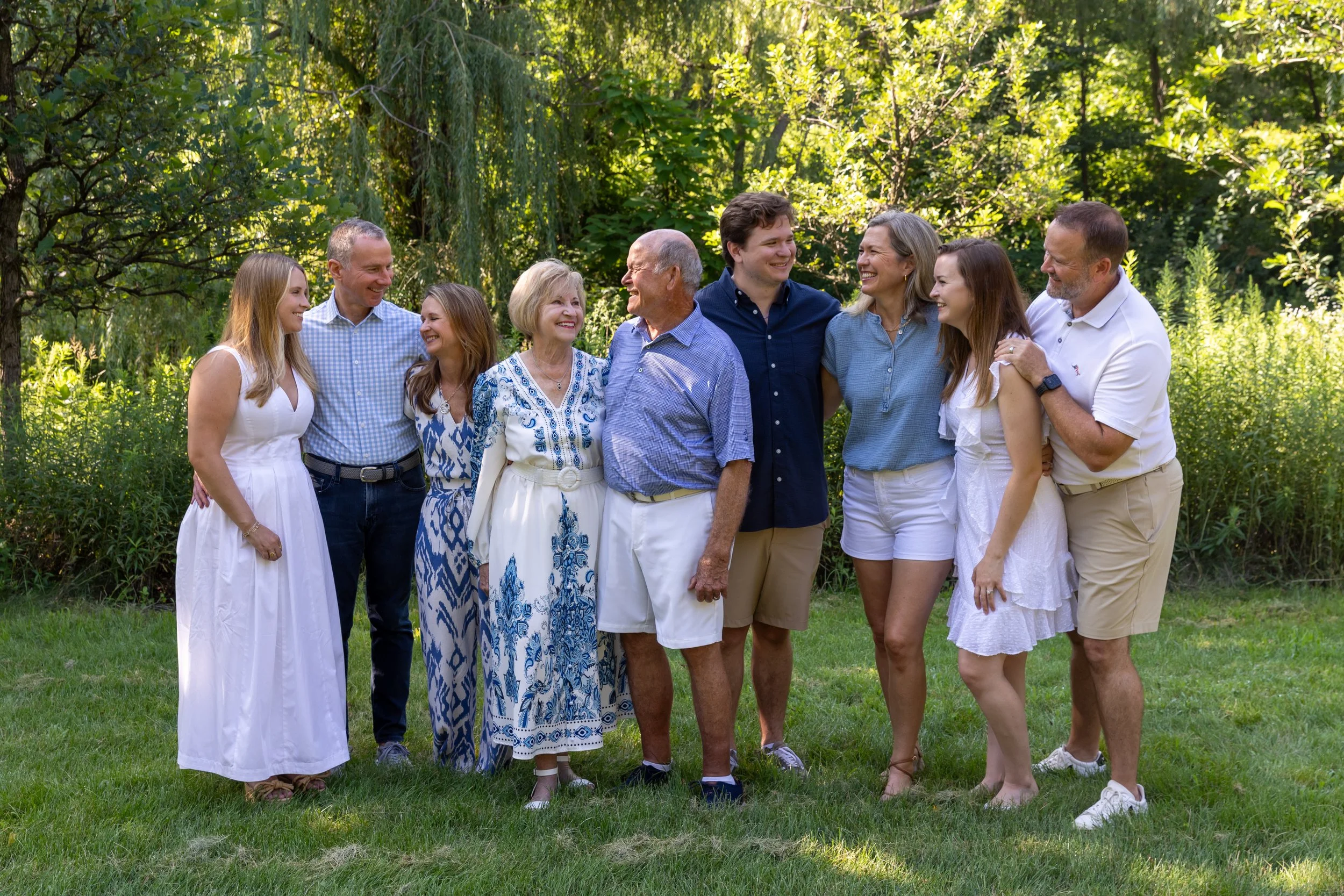 Three families stand together smiling and laughing at each other. The grandparents are in the middle with their two adult children and their families on either side of them. They are standing in front of a willow tree at Minnetonka Mills Park.