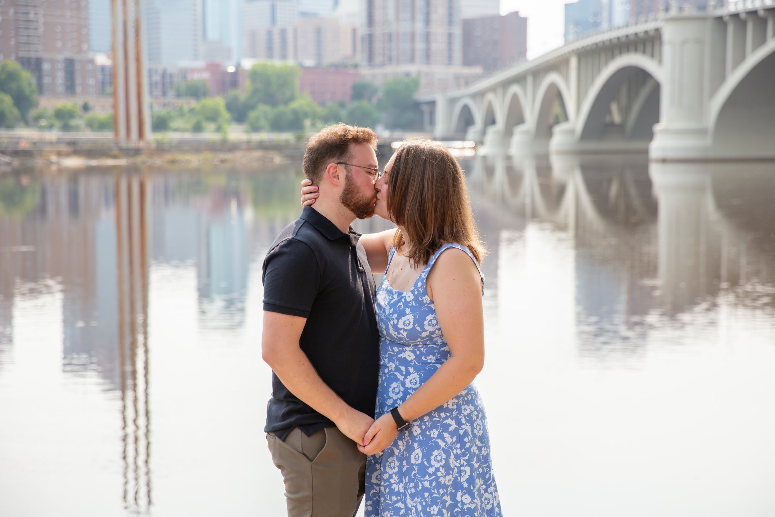 A couple, a man and woman, embraces each other kissing after he proposed to her in front of the falls and skyline at Water Power Park on St. Anthony Main.