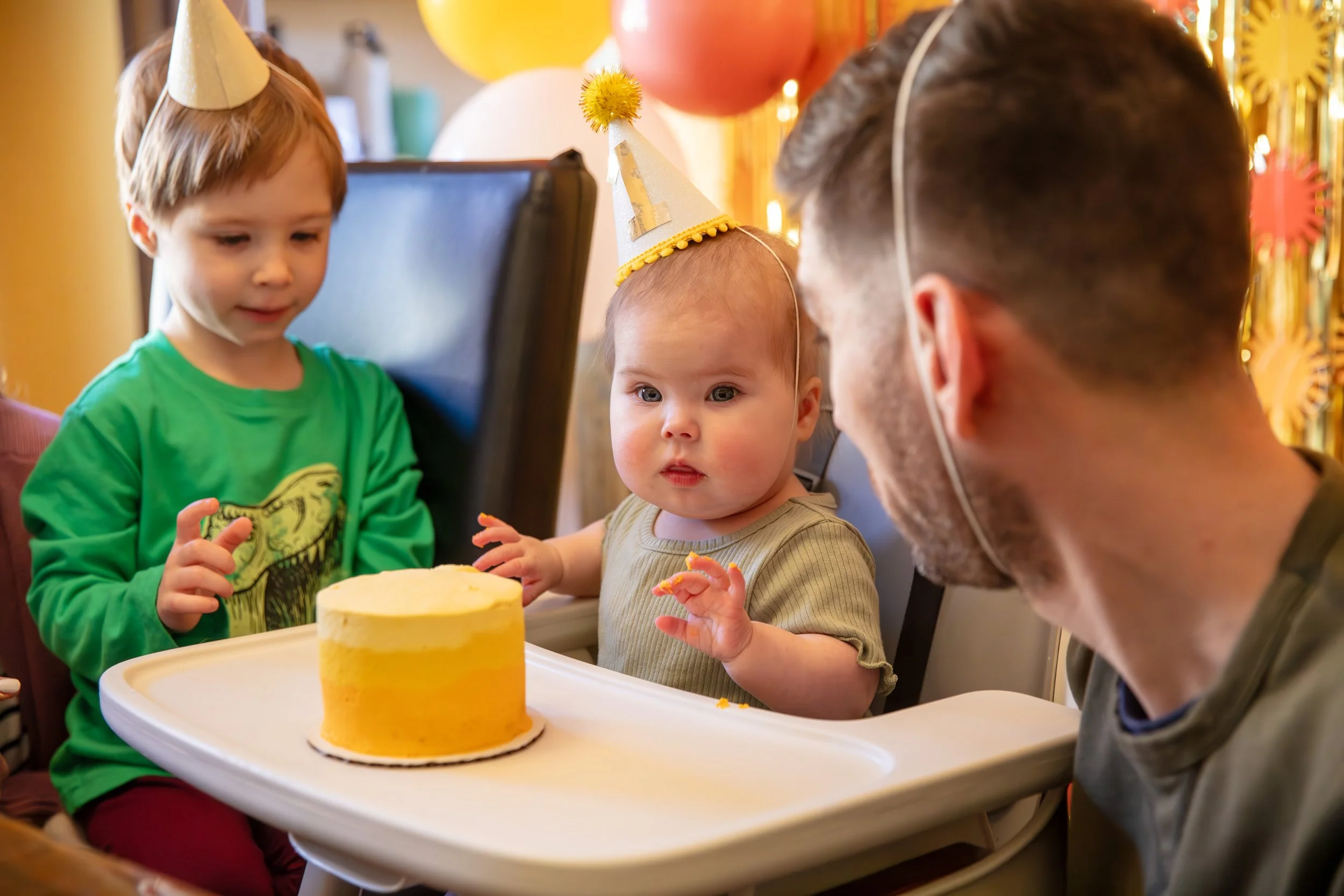 Minneapolis Cake Smash photographer - photo of a one year old baby in her highchair with a yellow frosted cake on the tray. She sits in front of birthday streamer and balloon decorations. Her father is on the right side of her making a funny face at 