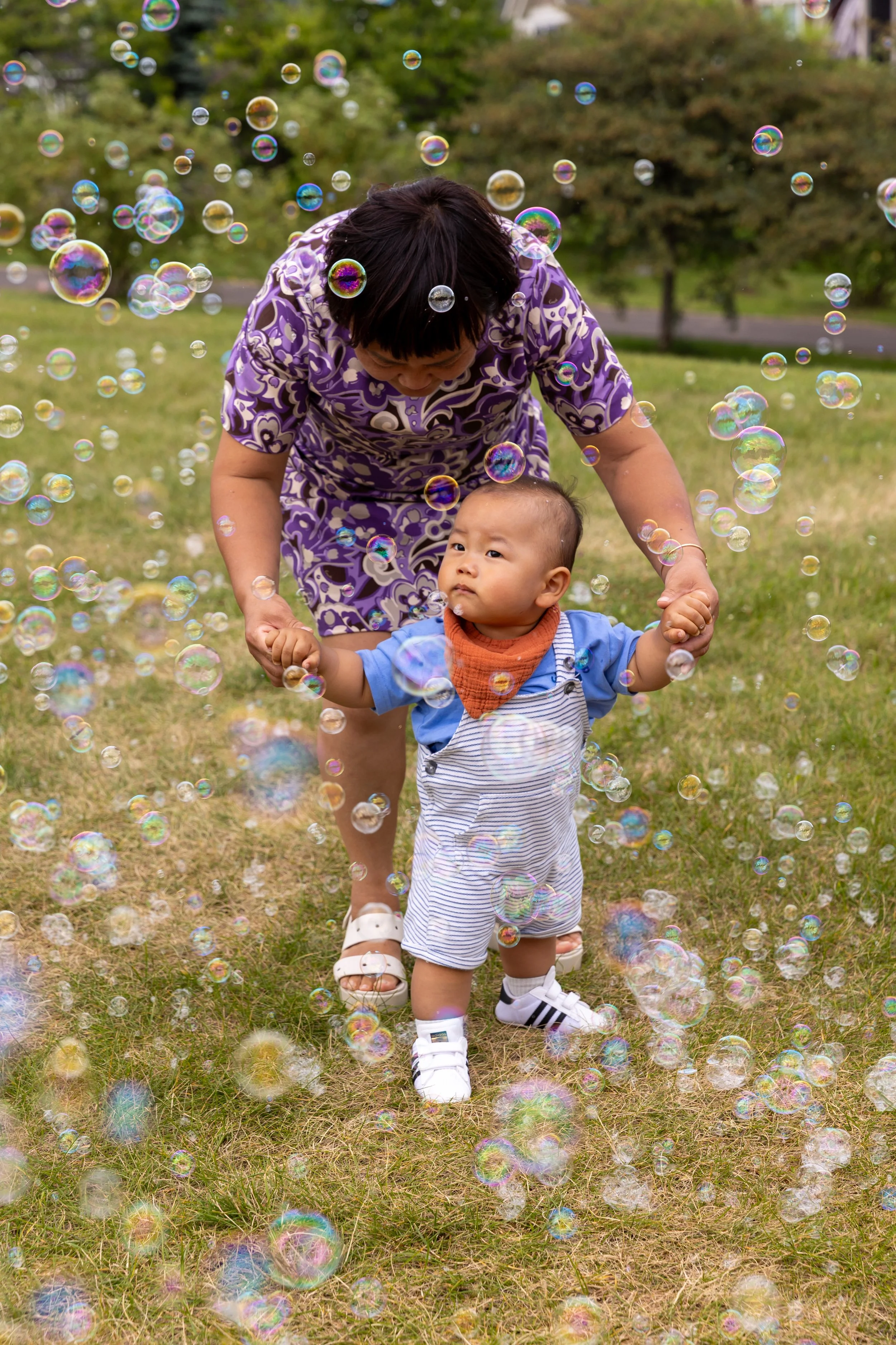 A one year old in white and blue striped overalls stands around bubbles blowing from a bubble machine in a St. Paul park his grandma in a purple dress holding his hands. 