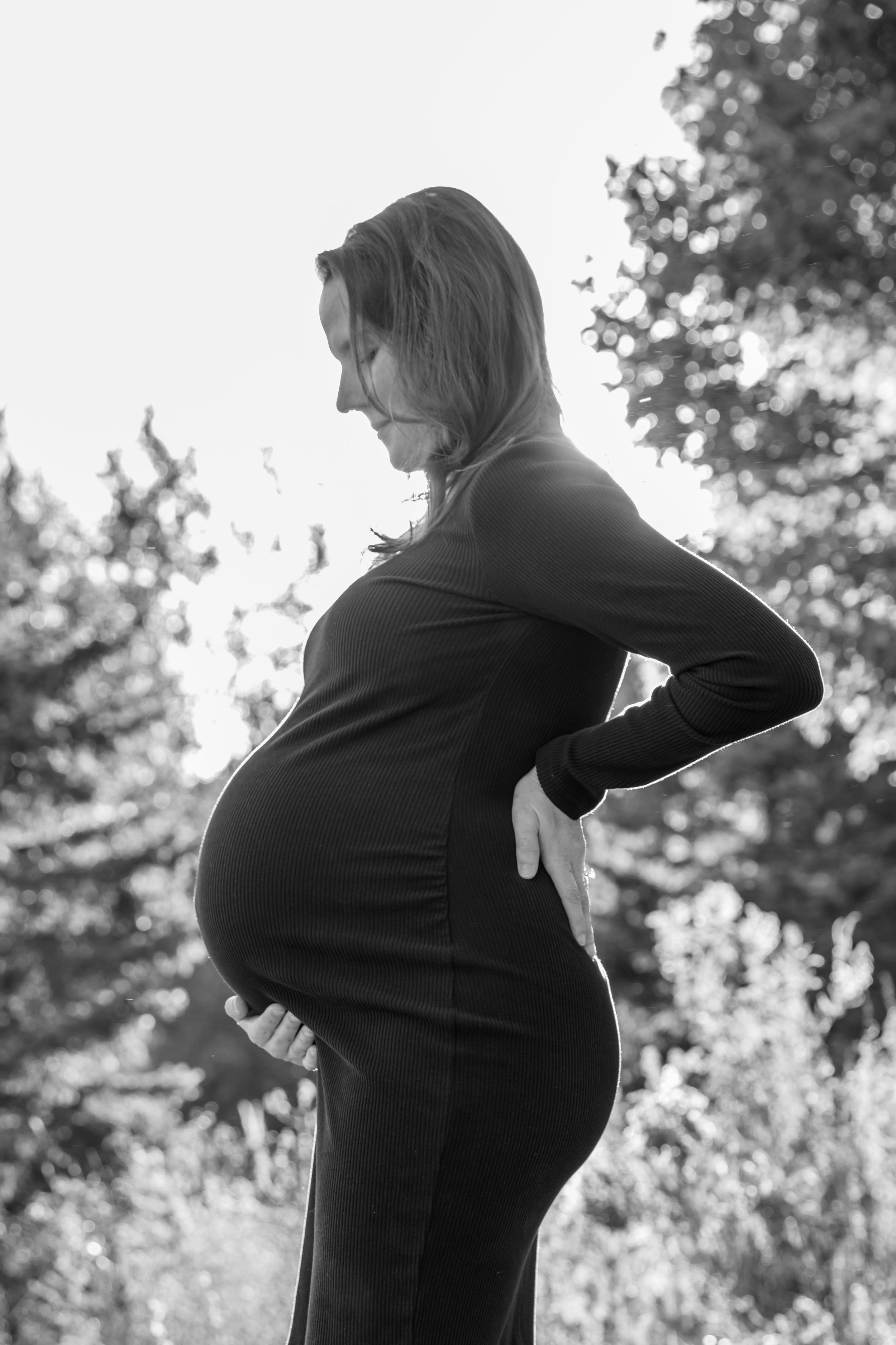 Maternity session, black and white silhouette of a profile of the expecting mother cradling her baby bump smiling down at it with the sun in the background.