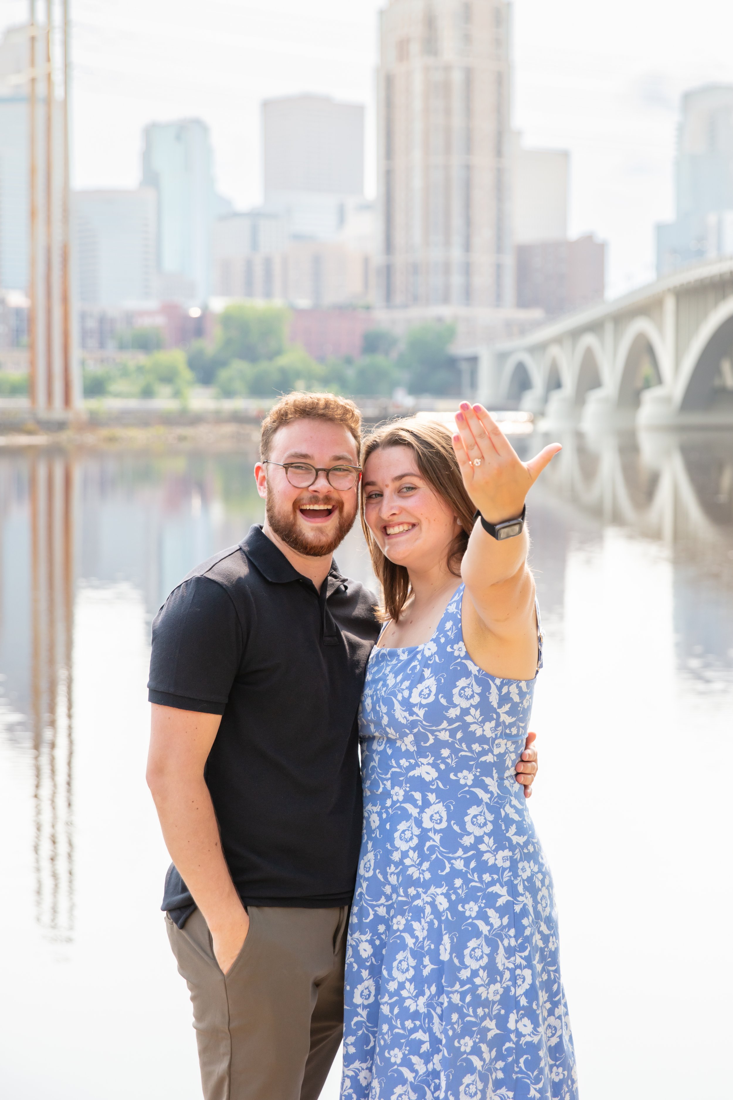 A couple, a man and woman, embrace each other and smile at the camera with her left hand in the air showing off her engagement ring after he proposed to her in front of the falls and skyline at Water Power Park on St. Anthony Main.