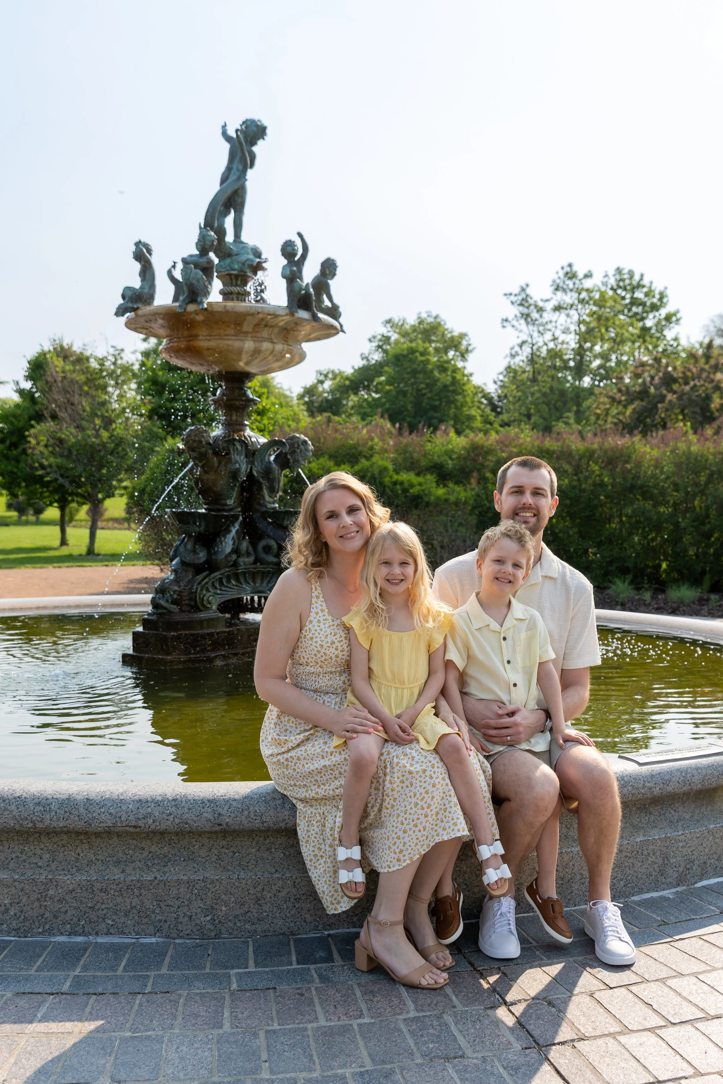 Minneapolis Family Photography May Mini Session- A mother and father sit on the edge of a water fountain with their twin son and daughter on their laps as they all smile at the camera together. They are at Lyndale Rose Garden in Minneapolis, MN. 