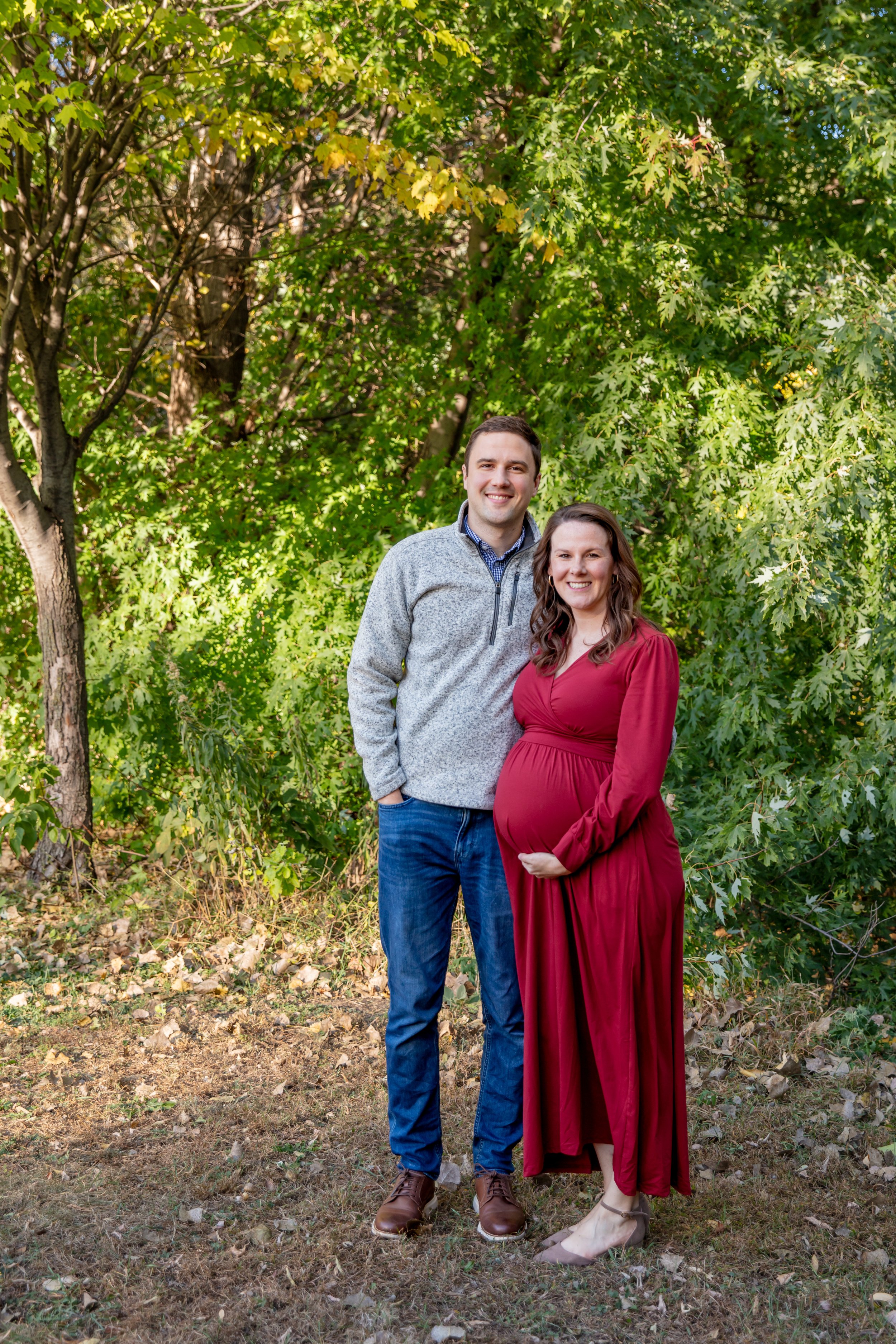 Maternity session, expecting mother and father-to-be embrace each other smiling at the camera in a Minneapolis park with fall foliage behind them.