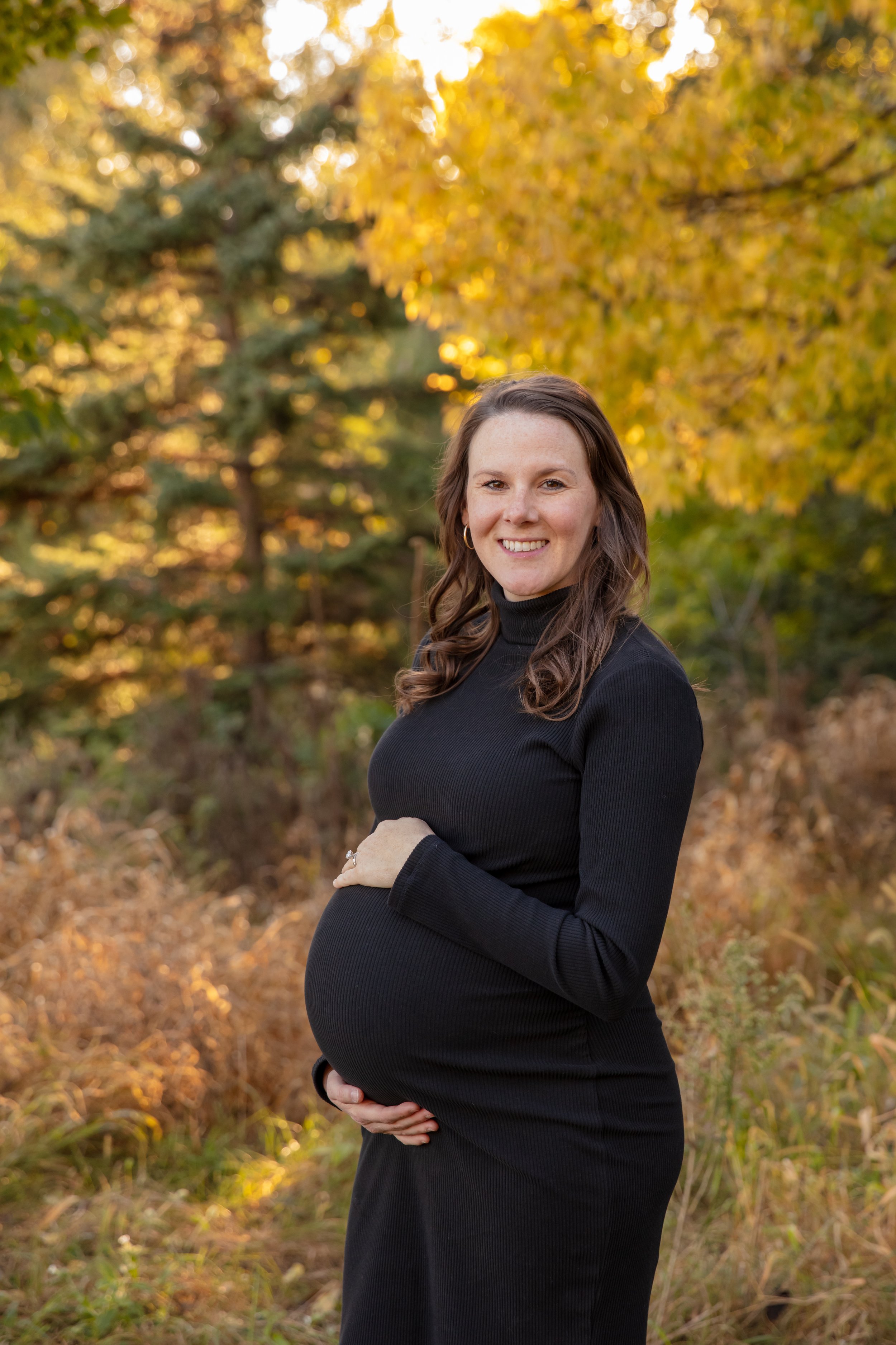 Maternity session, expecting mother cradles her baby bump smiling at the camera in a Minneapolis park with fall foliage behind them.