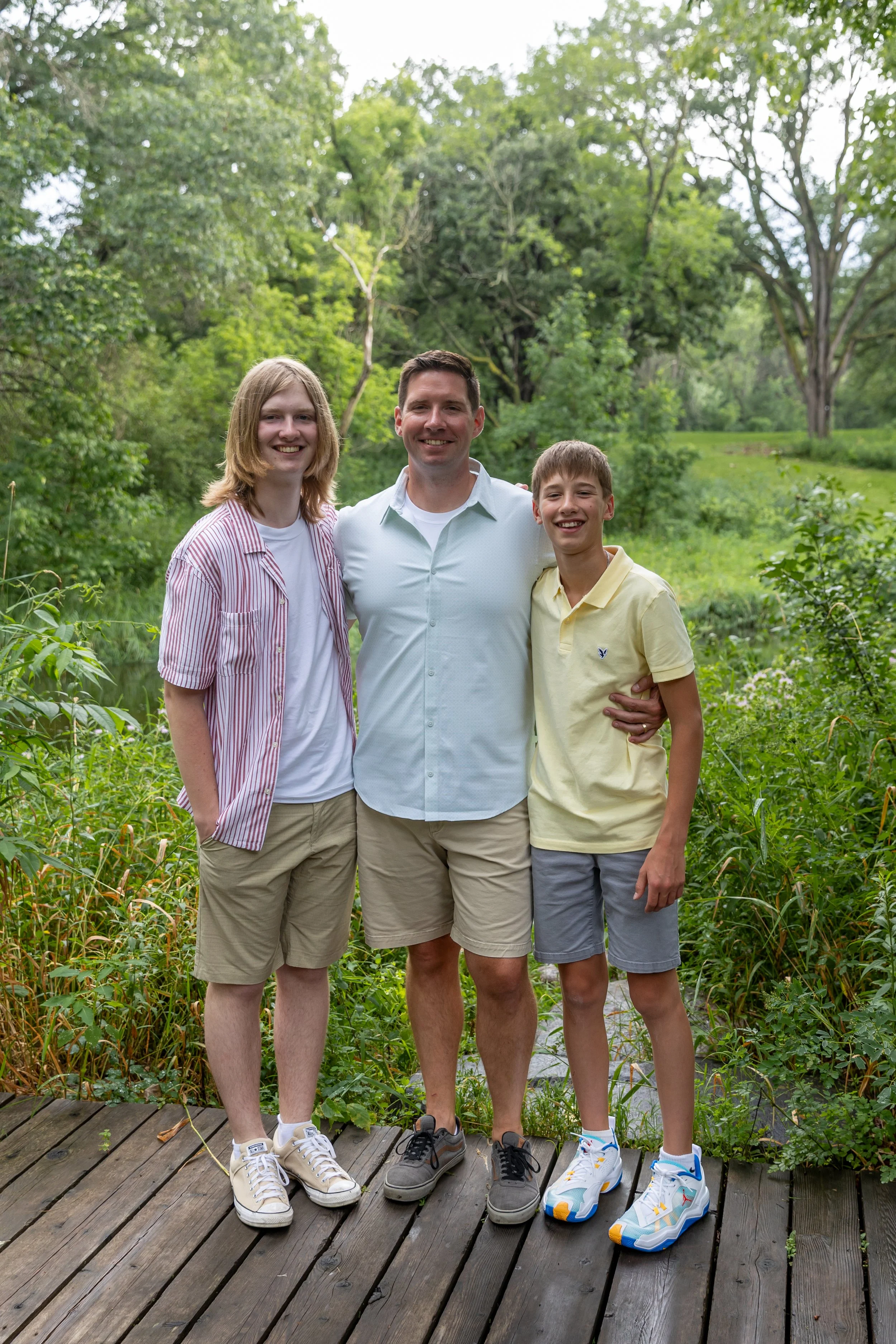 A father stands in the middle of her two teenage sons at Minnetonka Mills Park in Hopkins, MN while all smiling at the camera. 