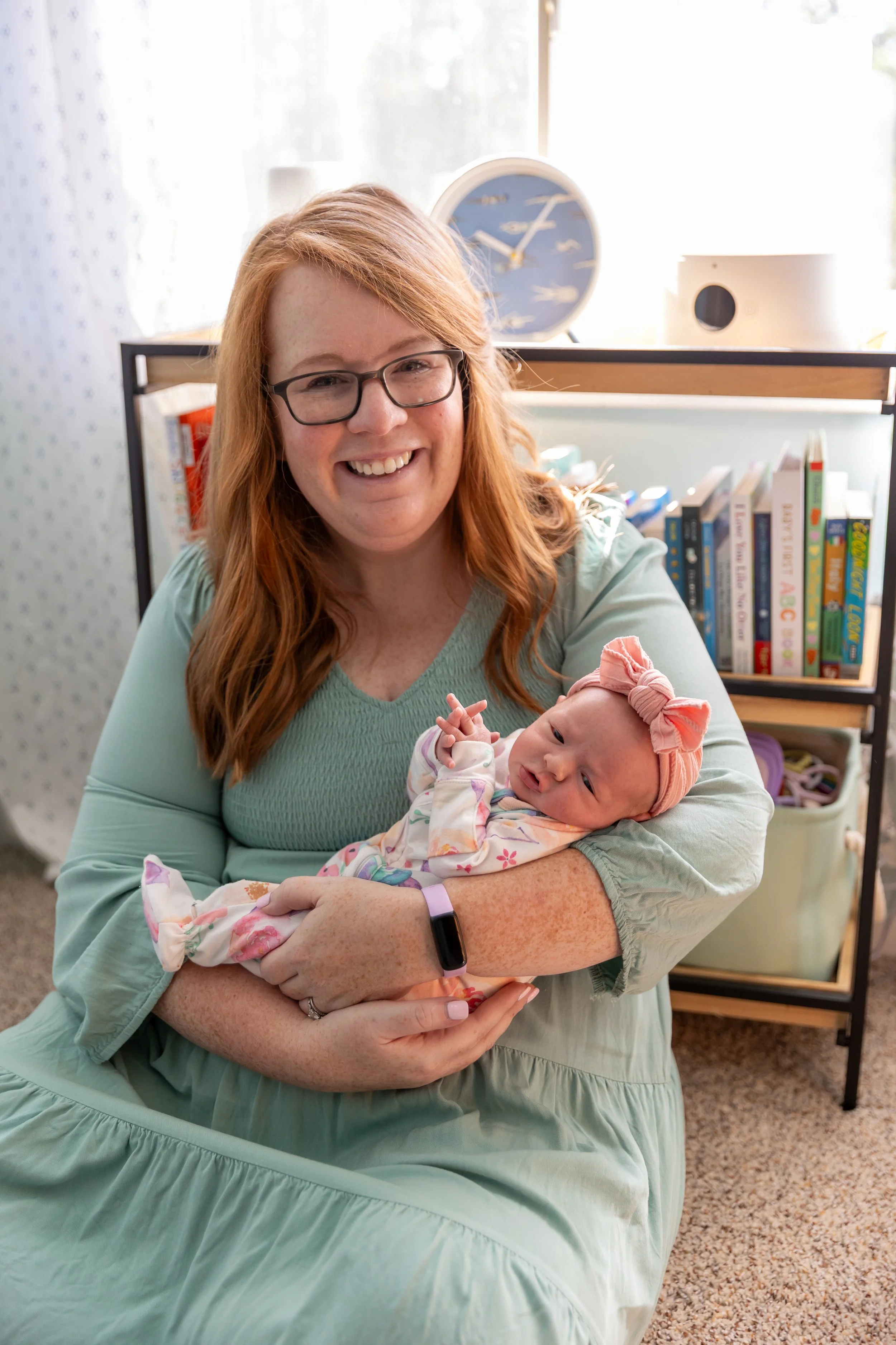 Minneapolis in-home newborn photography session - Mom holds her newborn baby in her arms as she smiles at the camera sitting on the floor in the nursery.