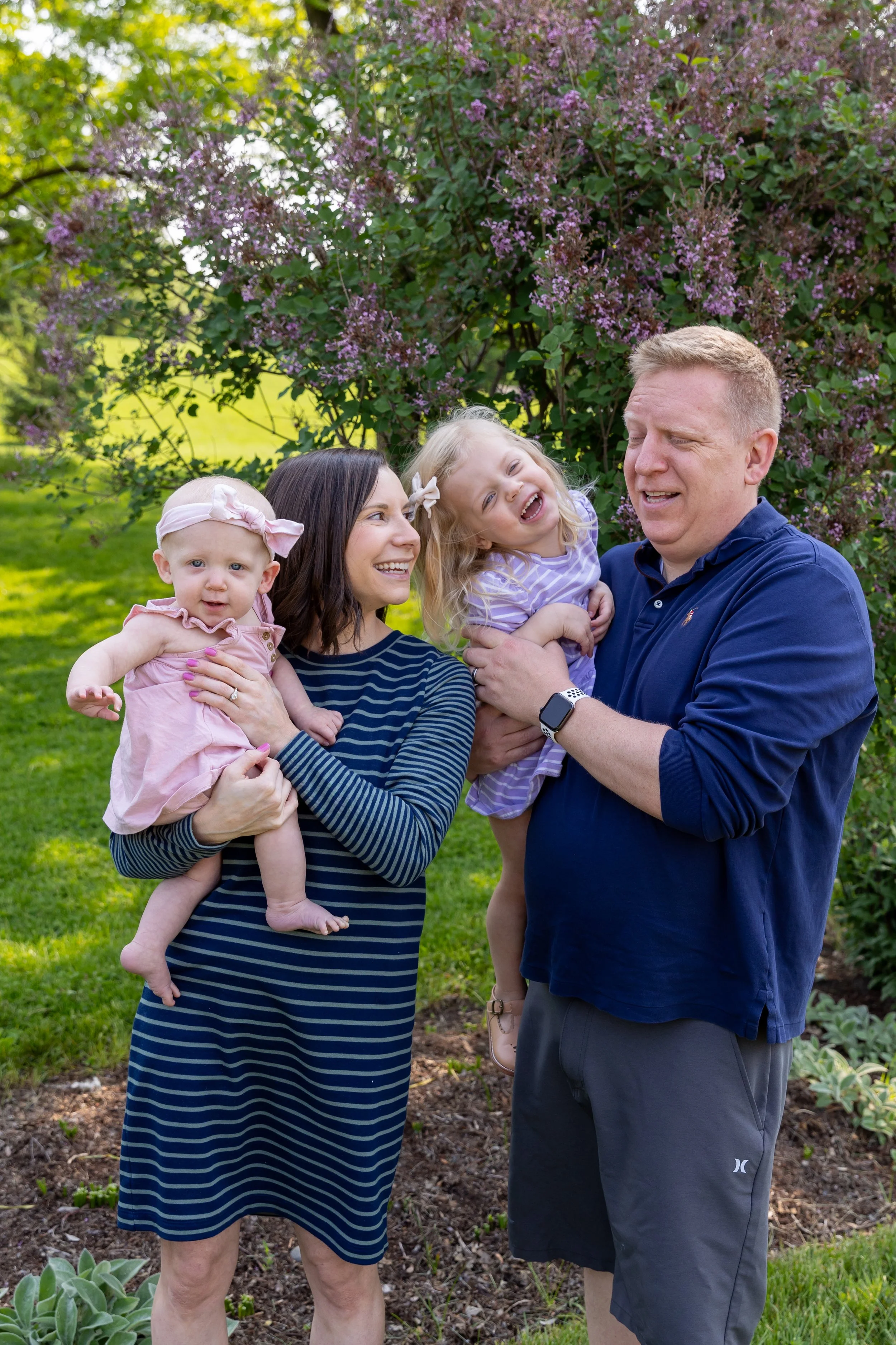Minneapolis family photography May Mini Session- A mother and father stand together holding their toddler and baby girls. The parents are tickling their daughters as they are all laughing at the camera. They are at Lyndale Rose Garden in Minneapolis,