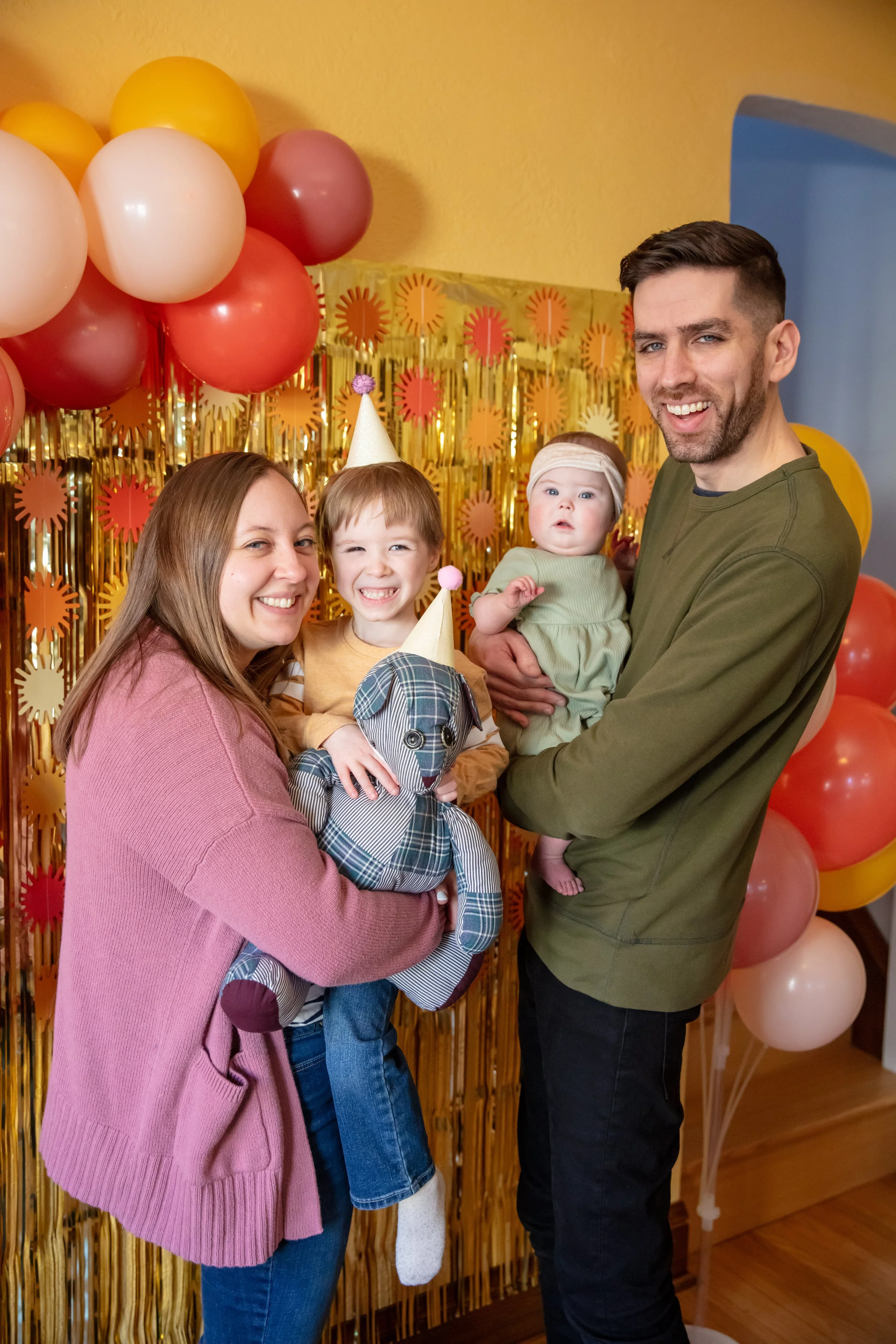 Minneapolis cake smash photographer - photo of a family of four. The mother and father hold their two children in between them as all four smile at the camera. They are surrounded by birthday streamers, balloons and decorations.