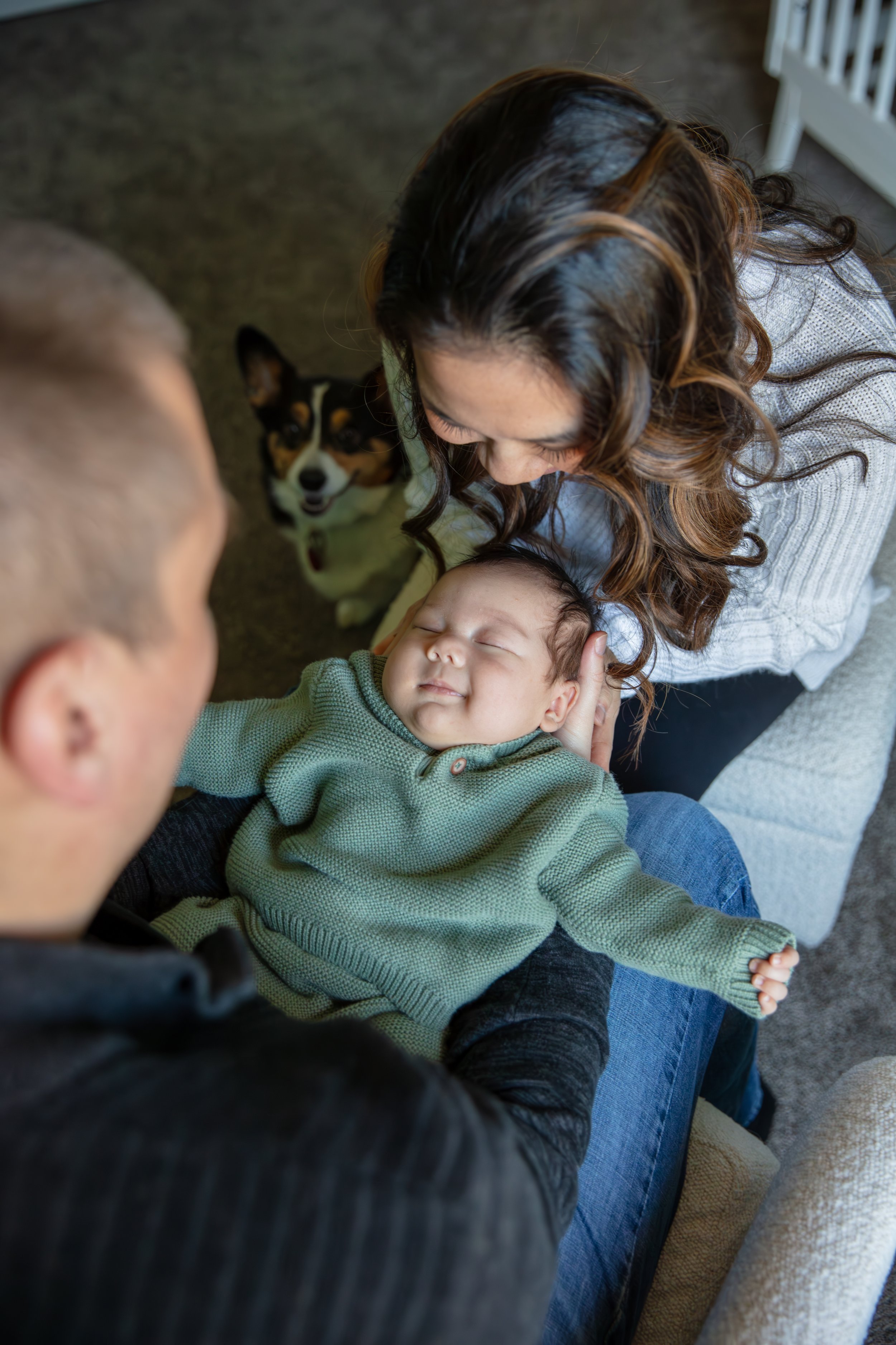 Newborn in-home session, close up over the father's shoulder holding his baby with mom holding the head from the other side smiling down at baby sleeping. Their corgi dog is in the background peeping up around the corner.