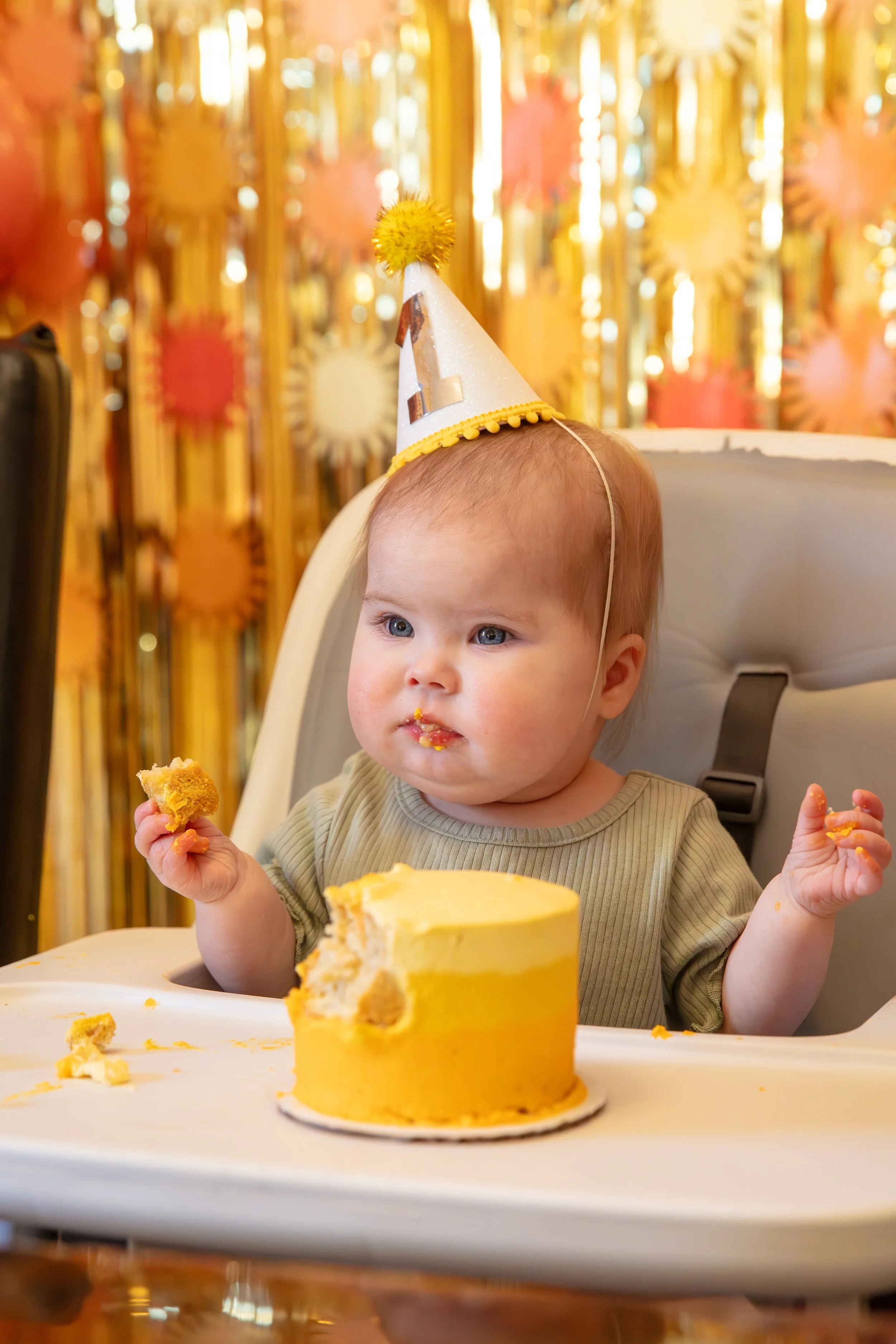 Minneapolis Cake Smash photographer - photo of a one year old baby in her highchair with a yellow frosted cake on the tray. She sits in front of birthday streamer and balloon decorations.  She has frosting on her face and hands.