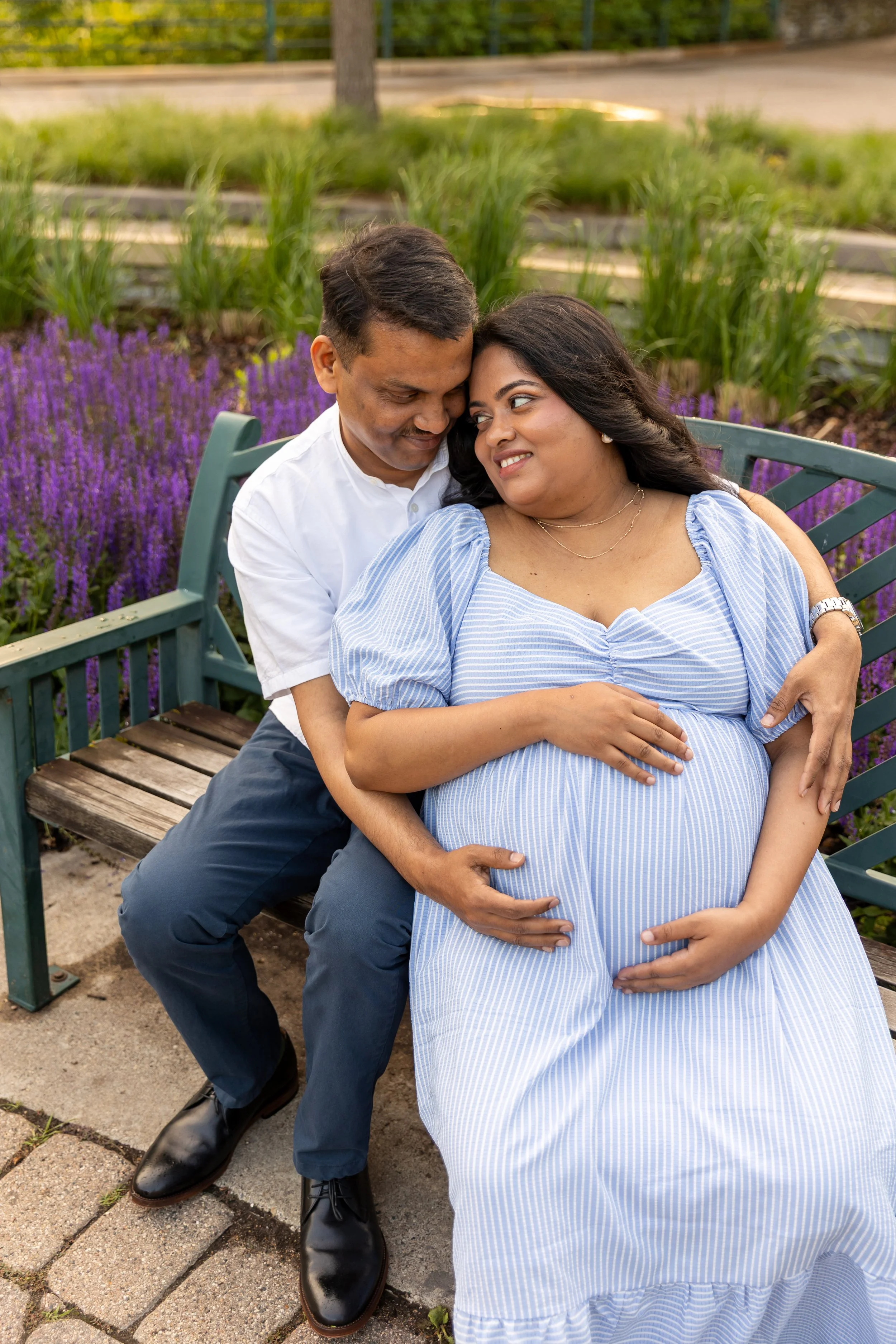 Maternity session - father and mother sit on a bench together while gazing lovingly at each other at Minnehaha Falls Park while holding the baby bump