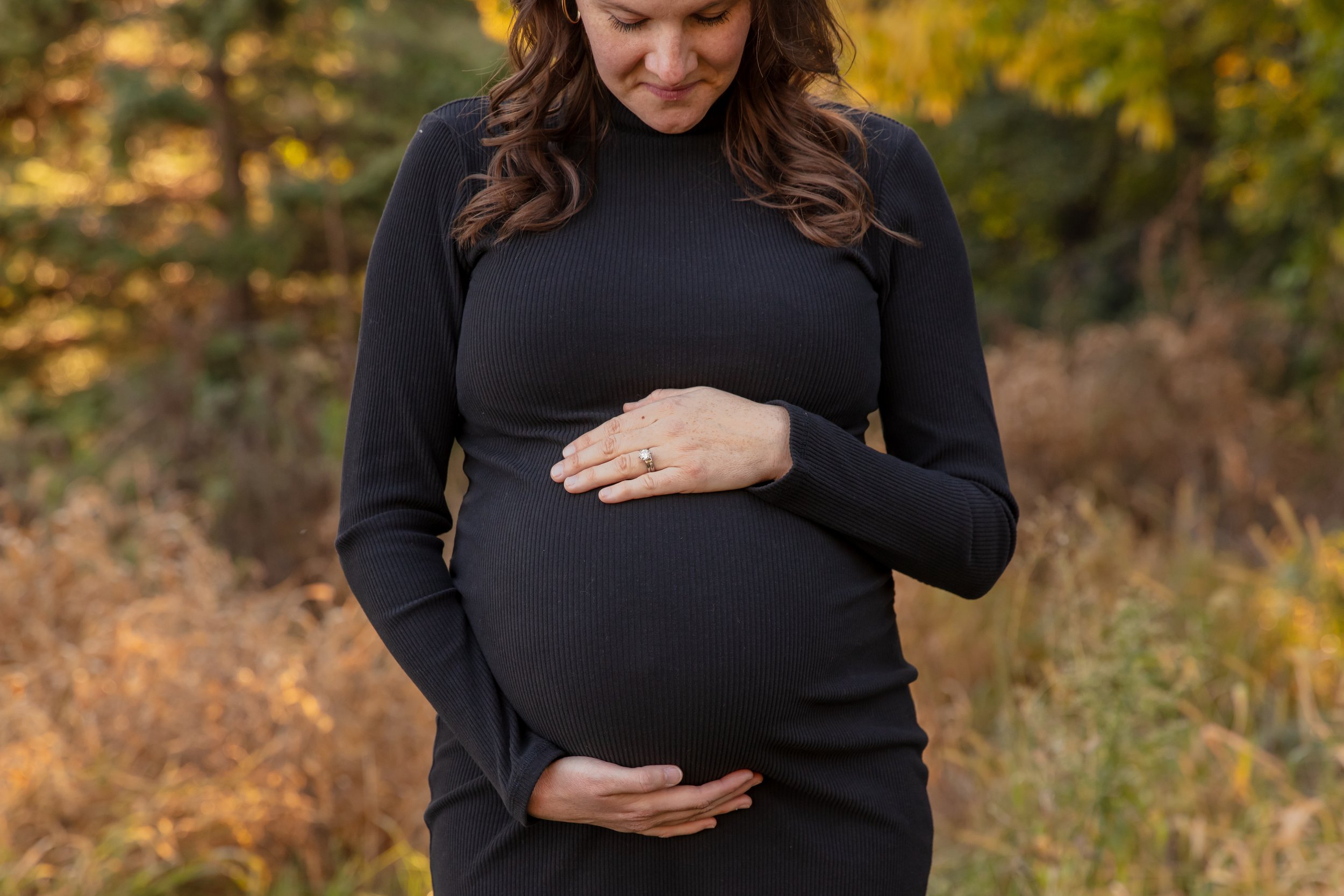 Maternity session, expecting mother cradles her baby bump smiling down at it in a Minneapolis park with fall foliage behind her.
