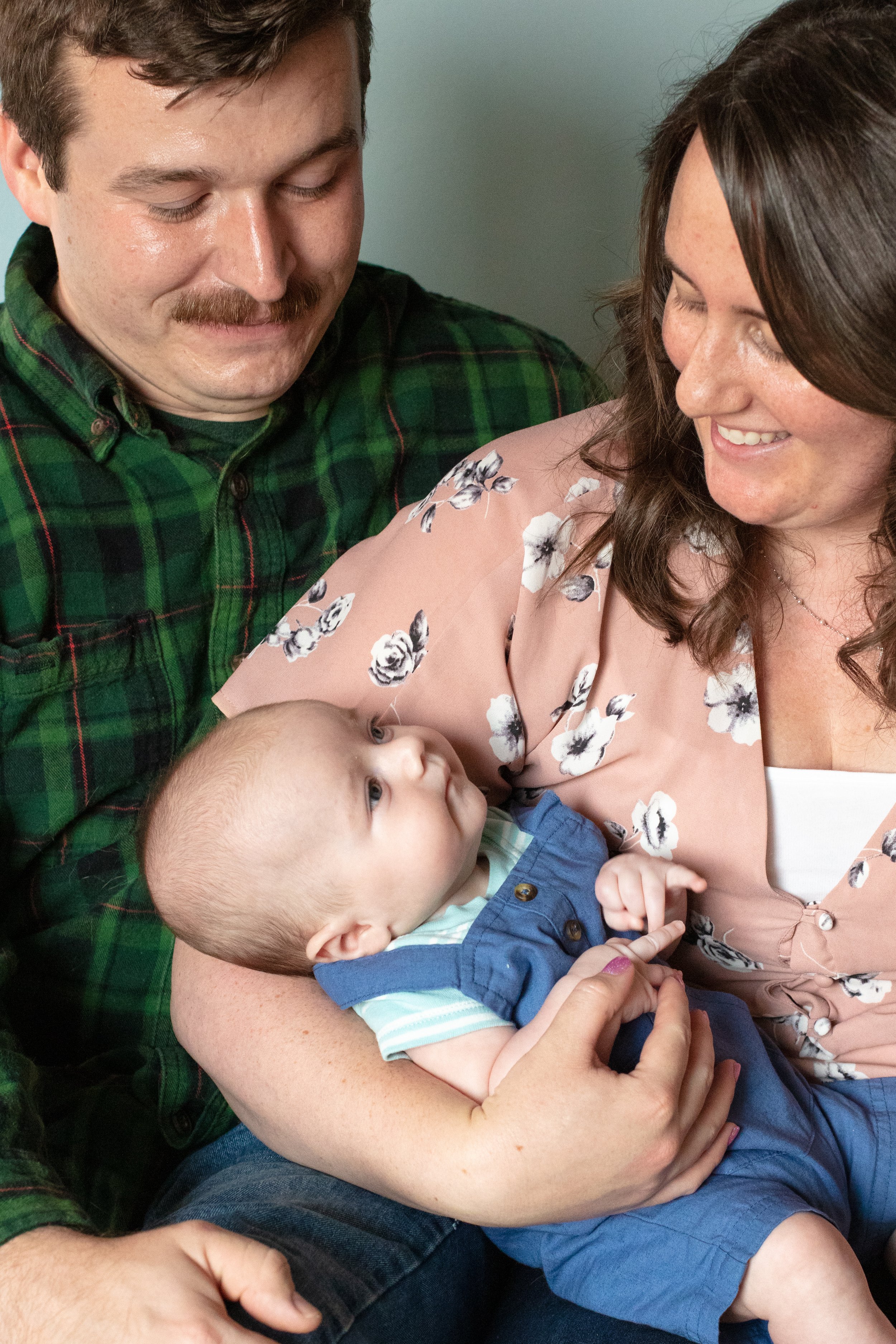 Up close image of parents sitting together while mom holds their baby. The baby gazes up at mom as both parents smile down at baby.