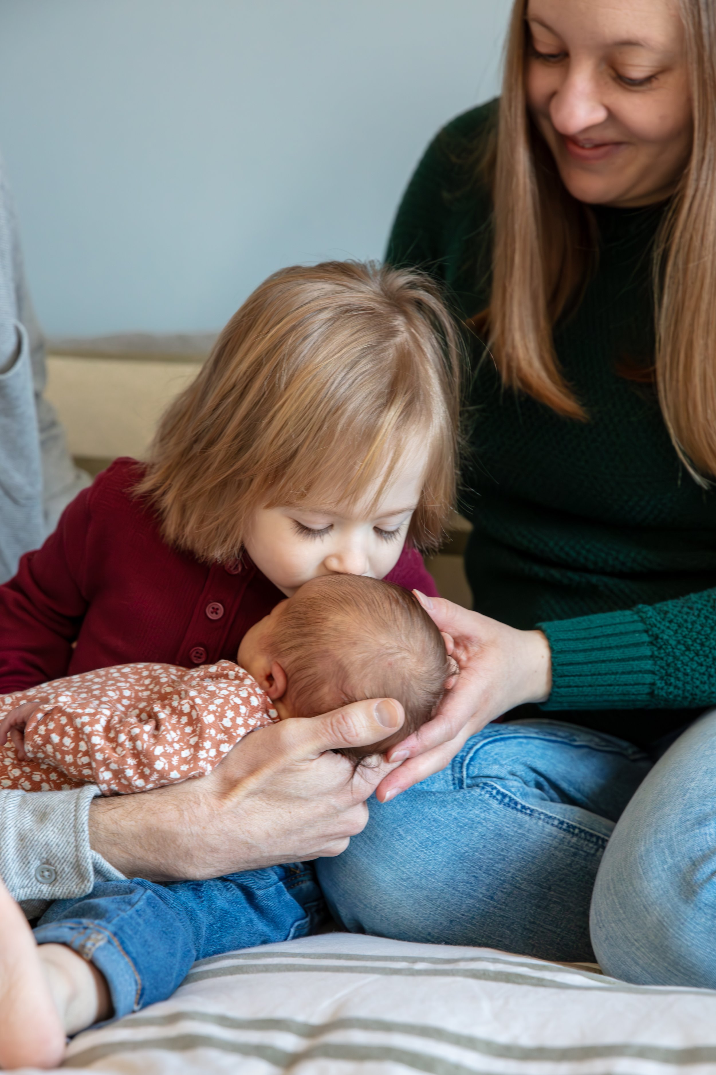 Up close of father and mother sitting on the bed (most of father is cropped out of the image) with their toddler between them as he leans over to his baby sister in his lap and kisses her forehead.