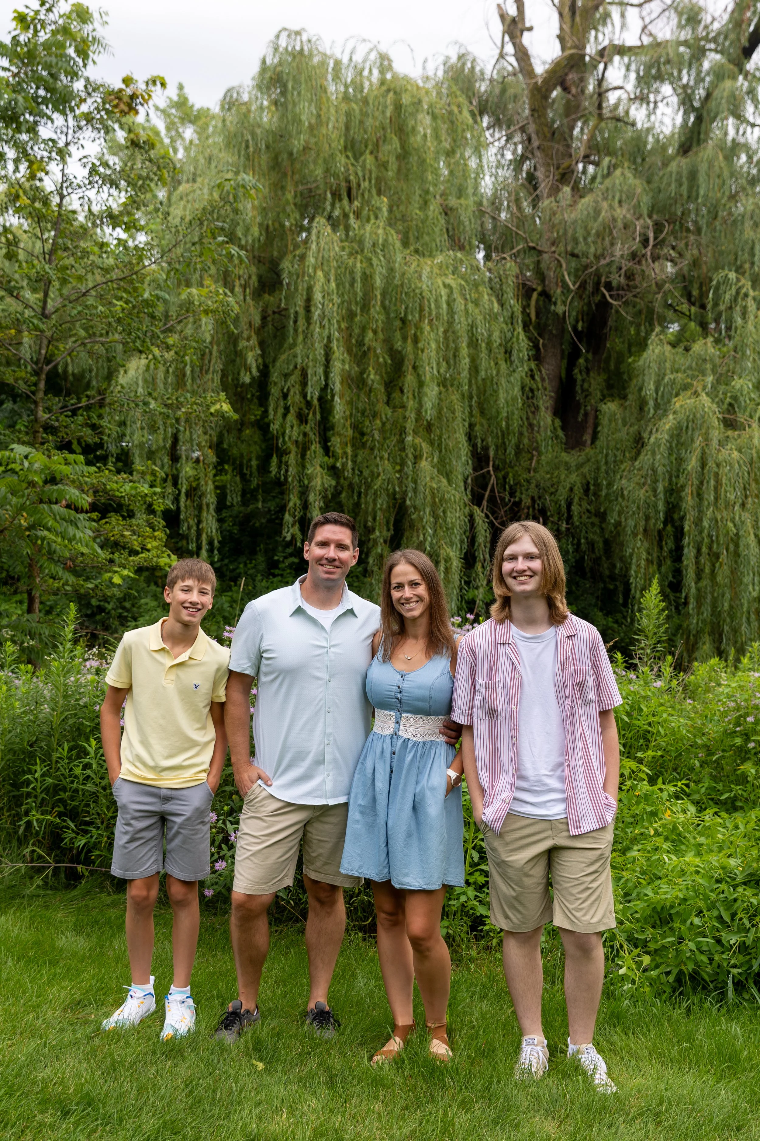 A family of four - a father and mother stand in the middle of their two teenage sons - in front of a willow tree at Minnetonka Mills Park in Hopkins, MN smiling at the camera. 